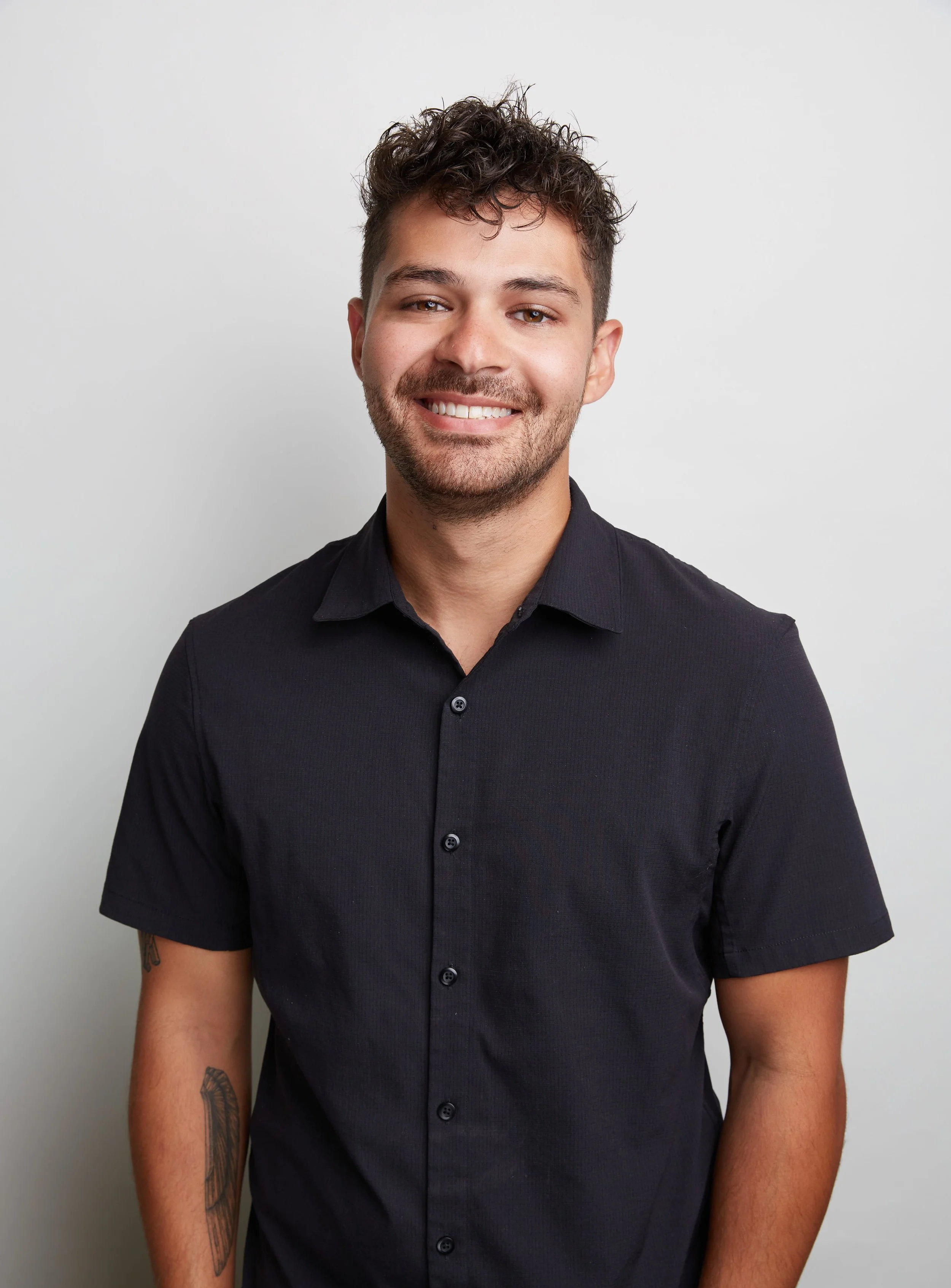 A young man with brown hair styled in curls, wearing a black button-up shirt, smiling and standing against a plain white background.