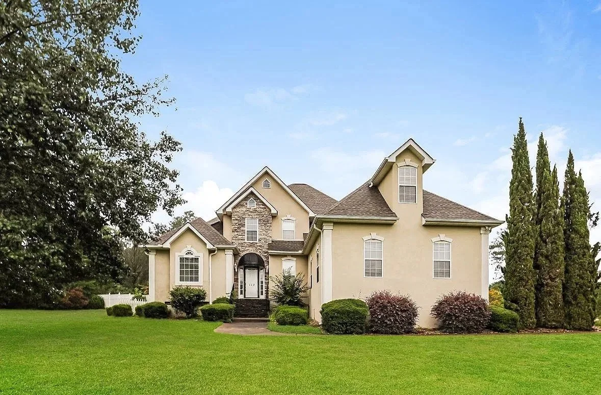 A large, beige two-story house with a dark brown roof and stone accents, surrounded by a well-kept lawn and tall trees.