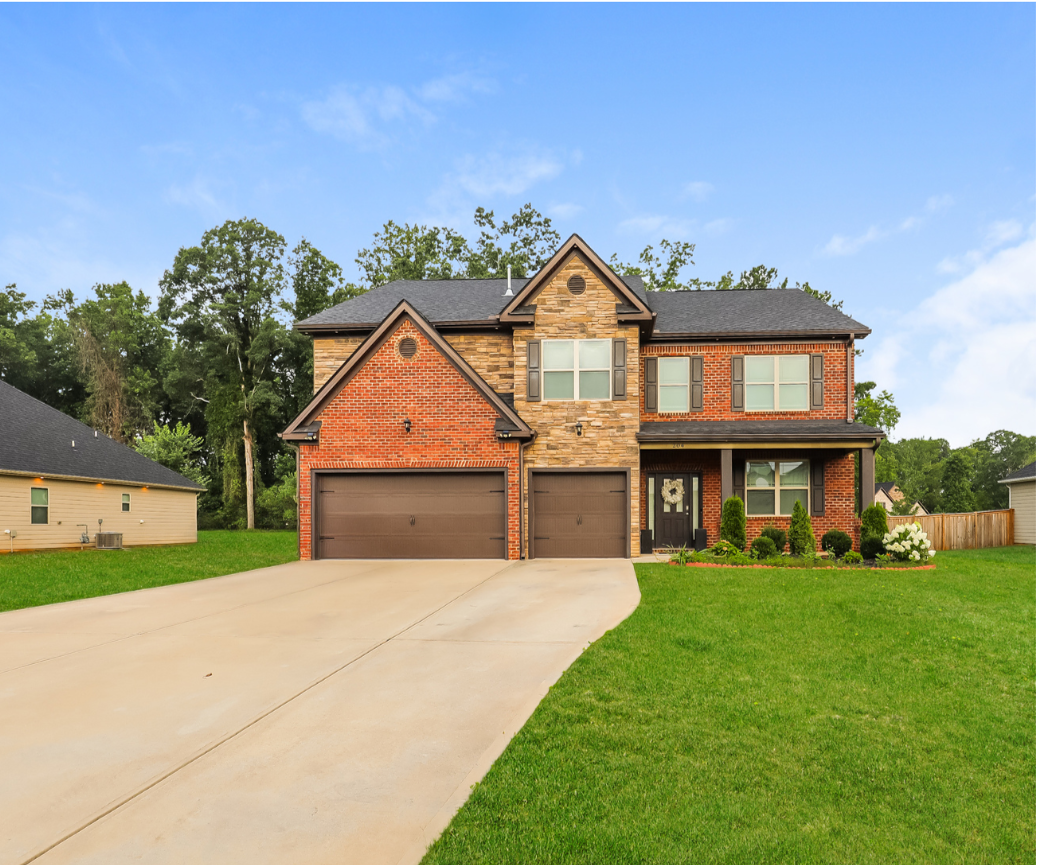 A two-story suburban house with a brick and stone exterior, a three-car garage, a concrete driveway, and a well-maintained lawn with shrubs and flowers.