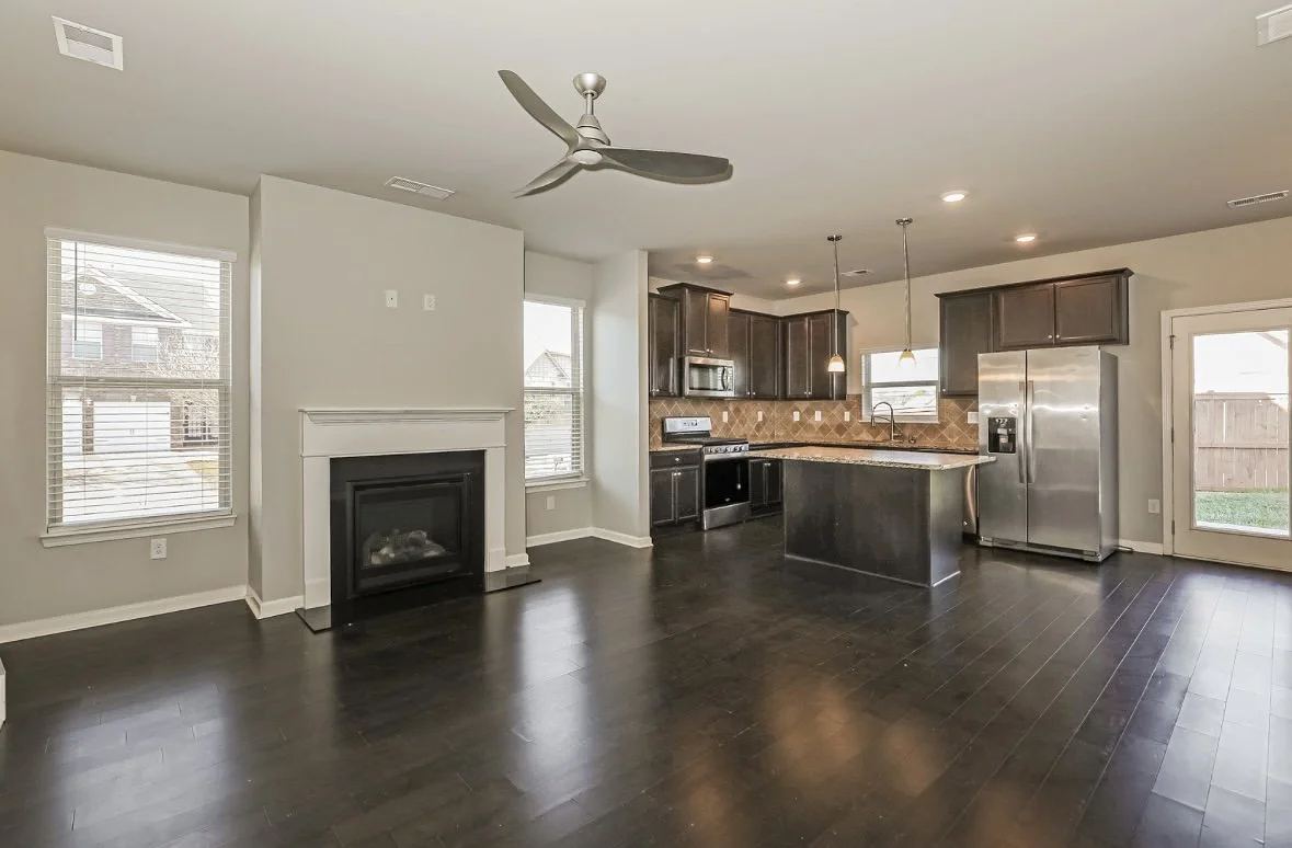 Empty living room and kitchen area with dark hardwood floors, gray walls, a fireplace, kitchen cabinets, stainless steel appliances, a kitchen island, and large windows.