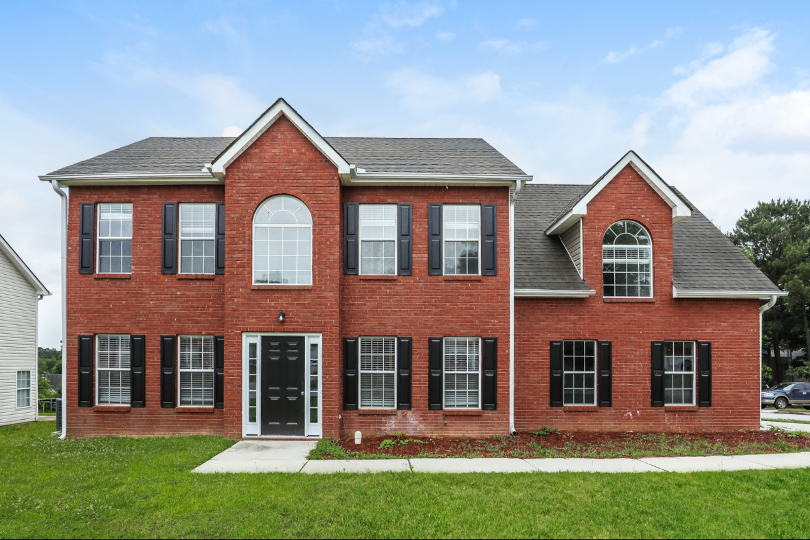 A two-story red brick house with black shutters and large arched and rectangular windows, surrounded by a green lawn.