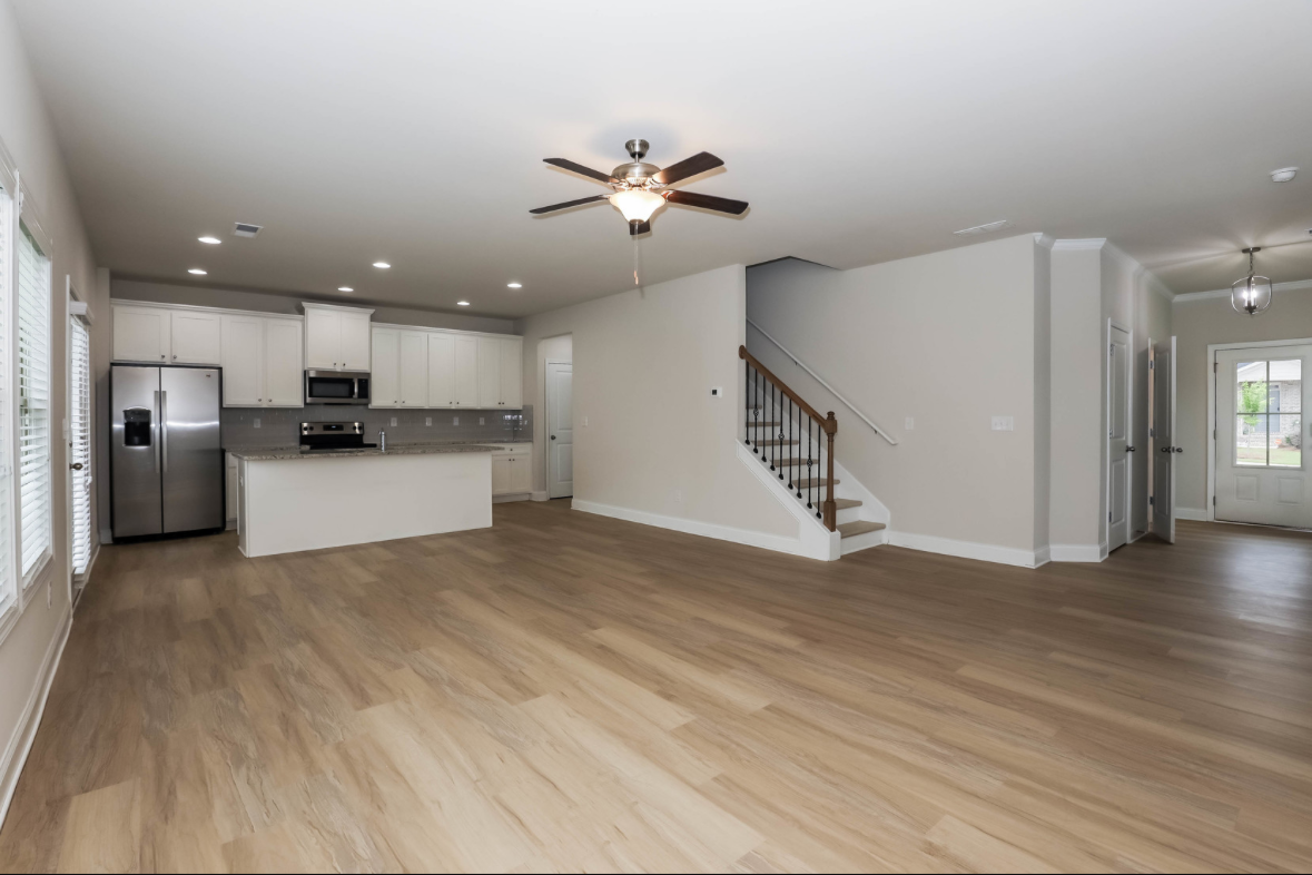 Open living space with hardwood floors, white kitchen cabinets, stainless steel refrigerator, and modern appliances, with staircase leading upstairs and a hallway with a front door in the background.