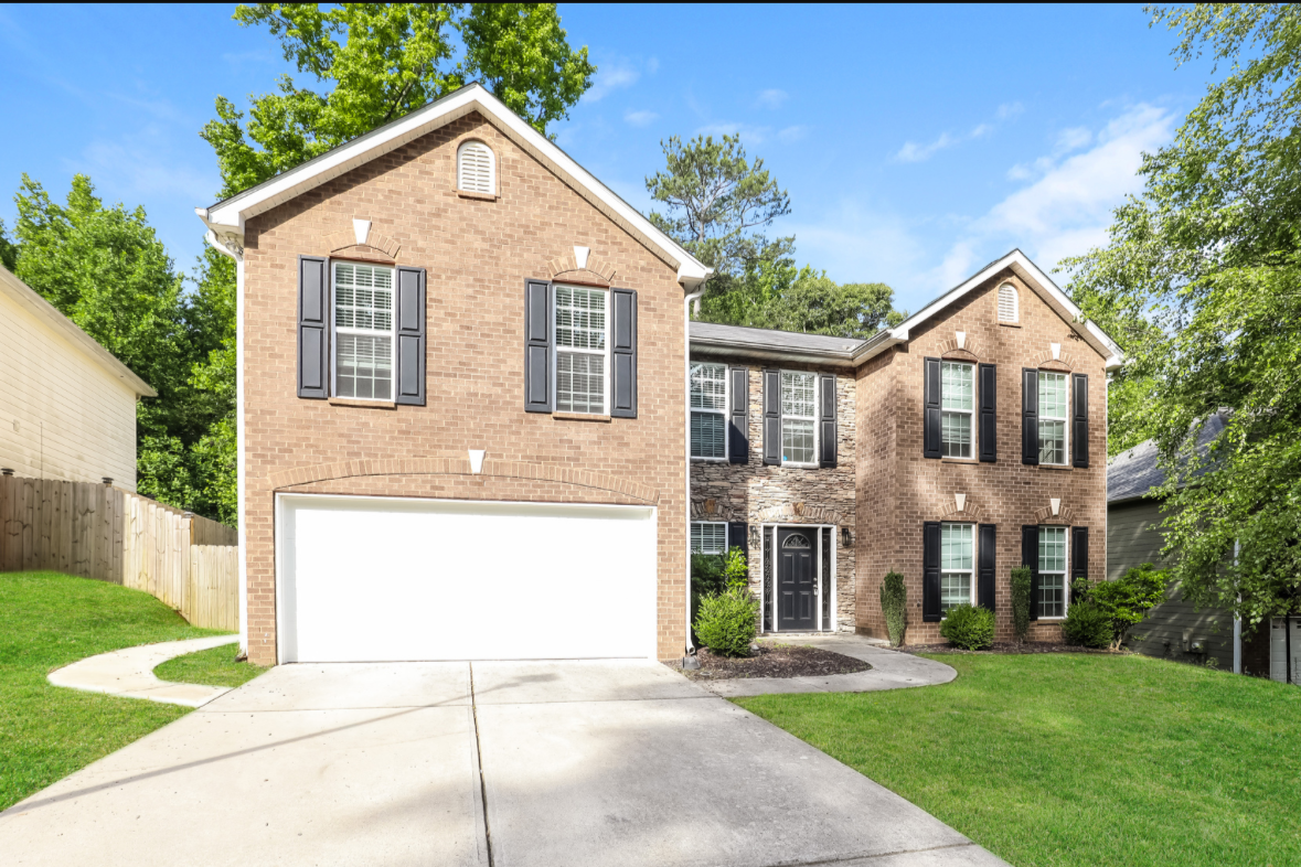 Front view of a two-story brick house with a two-car garage, black shutters, and a landscaped lawn with trees and shrubs.