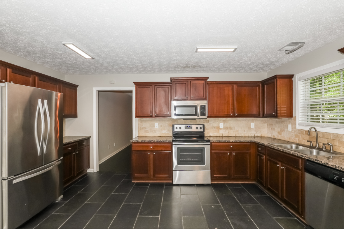Kitchen with dark wood cabinets, stainless steel appliances, granite countertops, beige tile backsplash, black tiled floor, and a window with blinds above the sink.