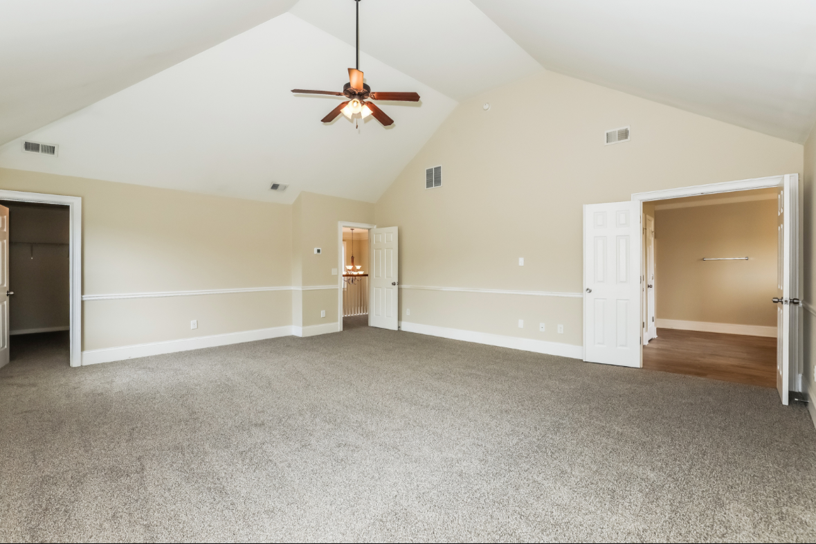 Empty beige living room with carpeted floor, vaulted ceiling, ceiling fan, and multiple open doors leading to closets and other rooms.