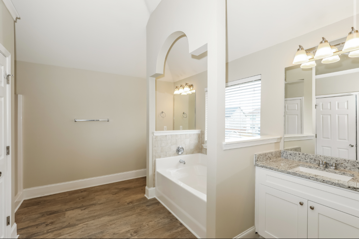 Bathroom with a bathtub, granite countertop vanity, large mirror, window with blinds, and beige walls.