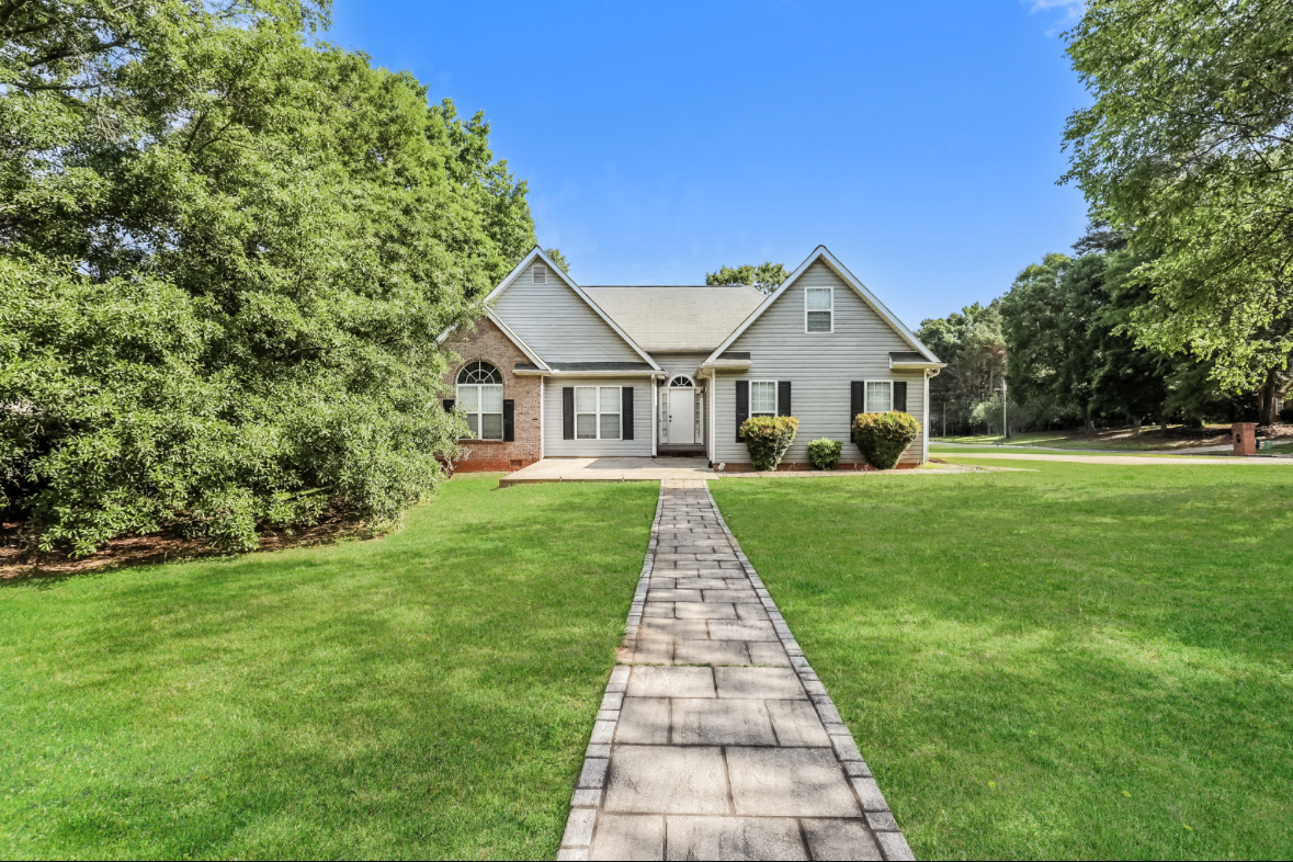 Front view of a house with a stone walkway leading to the front door, surrounded by green lawn and trees under a blue sky.