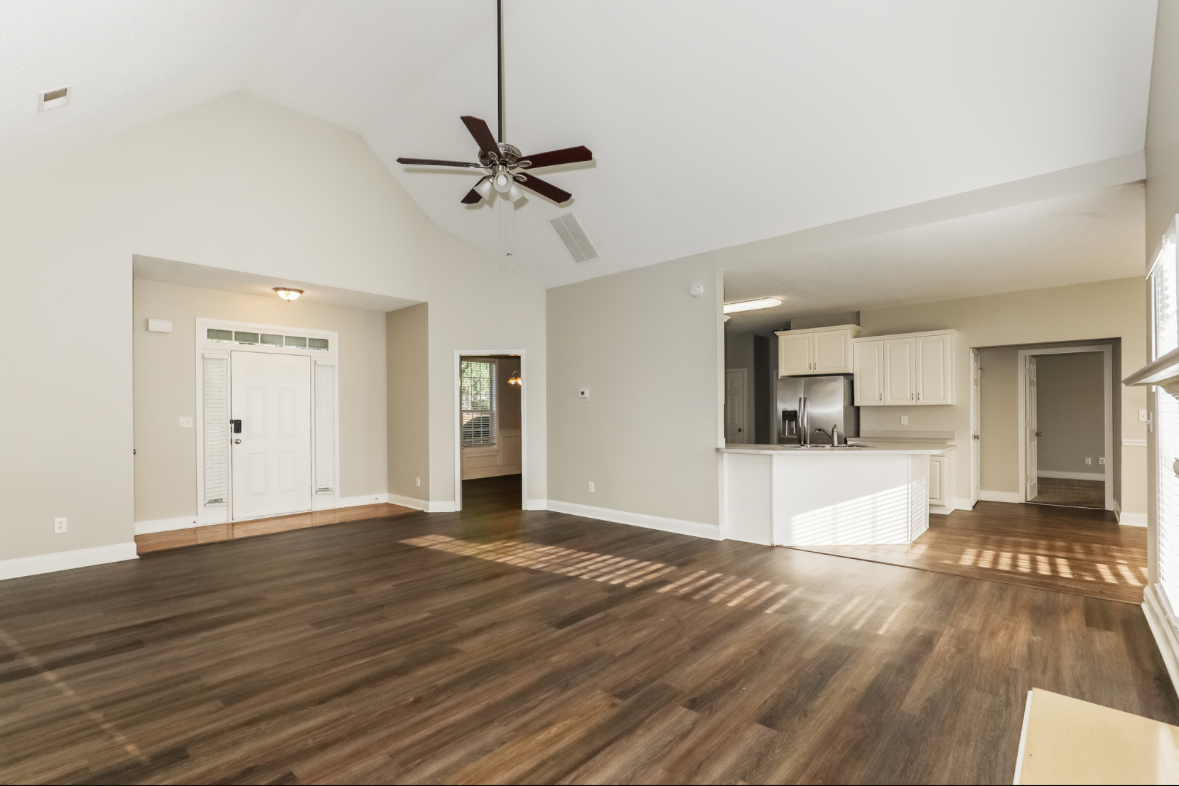 Empty living room with vaulted ceiling, ceiling fan, hardwood floors, white walls, kitchen with white cabinets and stainless steel appliances.