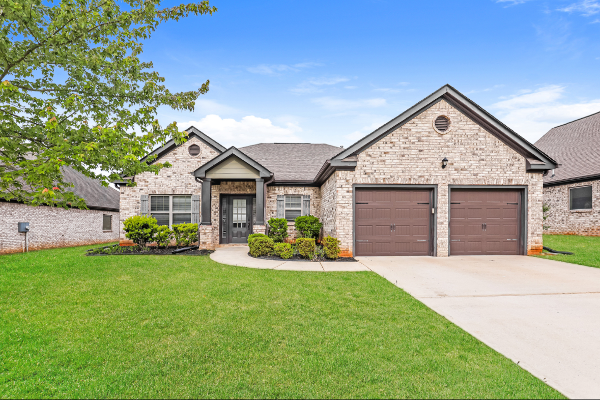 Front view of a brick house with a double garage, a green lawn, a walkway, and a tree on the left under a partly cloudy sky.