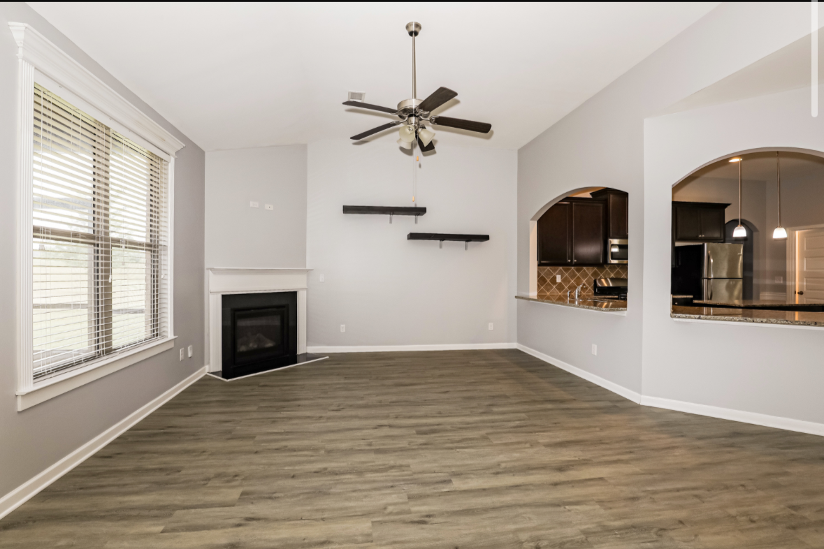 Empty living room with gray walls, wood flooring, large window with blinds, a white fireplace, ceiling fan, and open kitchen with dark cabinets and granite countertops