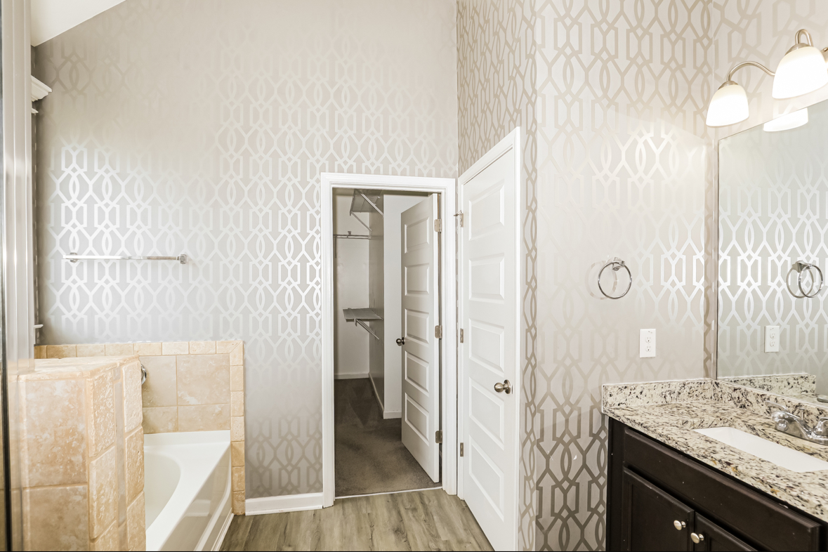 Bathroom with patterned wallpaper, granite countertop vanity with dark cabinets, large mirror, two wall-mounted lights, a bathtub with beige tile surround, and an open door leading to a walk-in closet with shelves and hanging space.