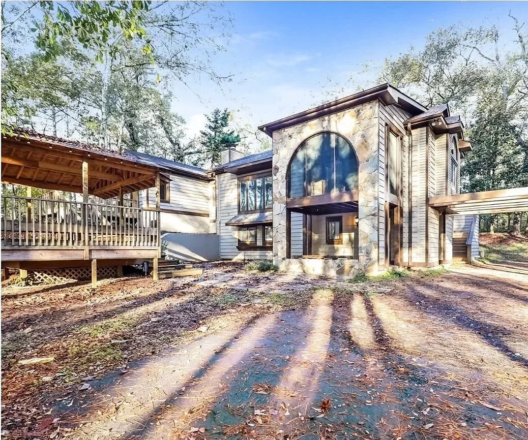 Large modern house with a stone façade and large arched window, surrounded by trees, with a wooden deck on the left, on a dirt ground with fallen leaves.Photographed by Branded by MDM 