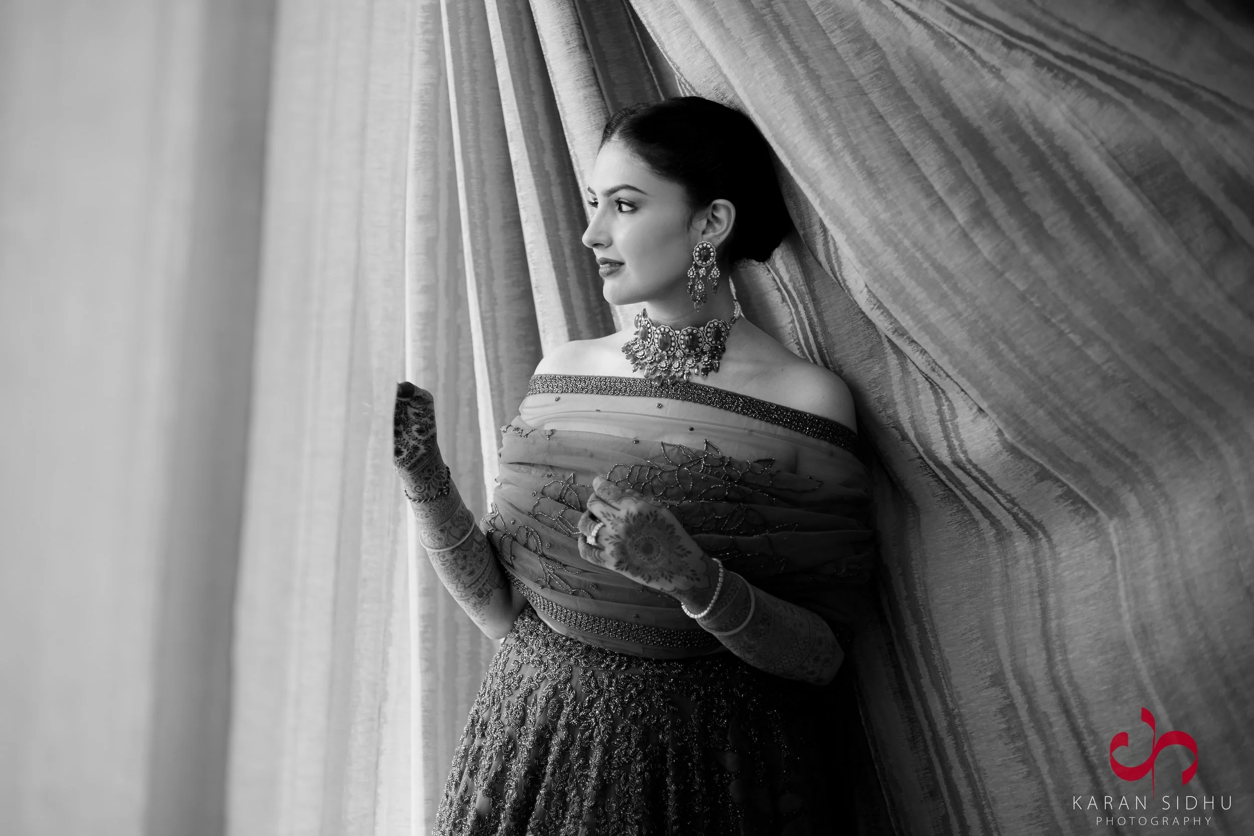 A woman dressed in traditional Indian attire, wearing jewelry and henna on her hands, looking to the side, leaning against curtains.