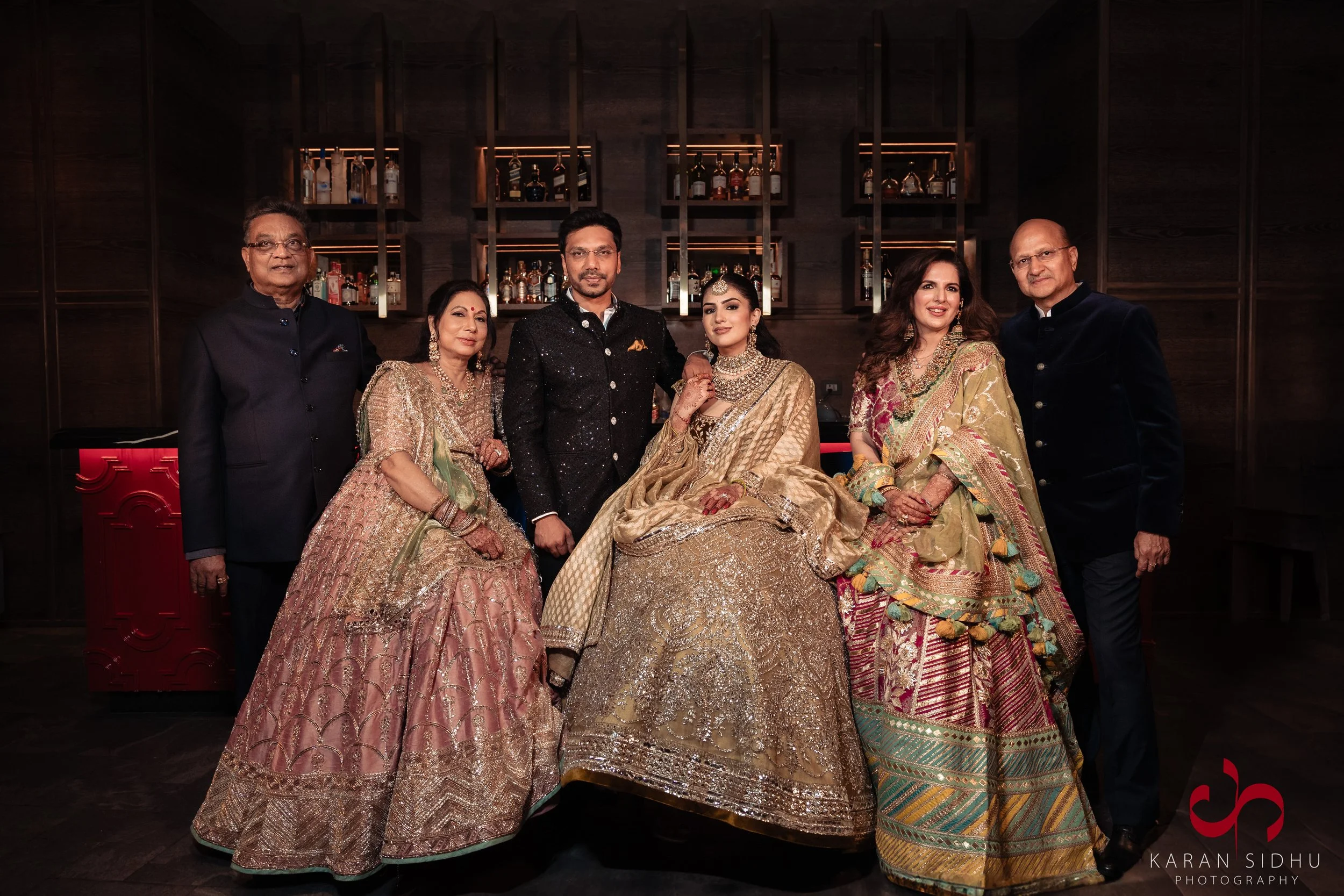Group of seven people dressed in traditional Indian attire posing in a dimly lit bar or lounge with bottles of liquor on shelves behind them.