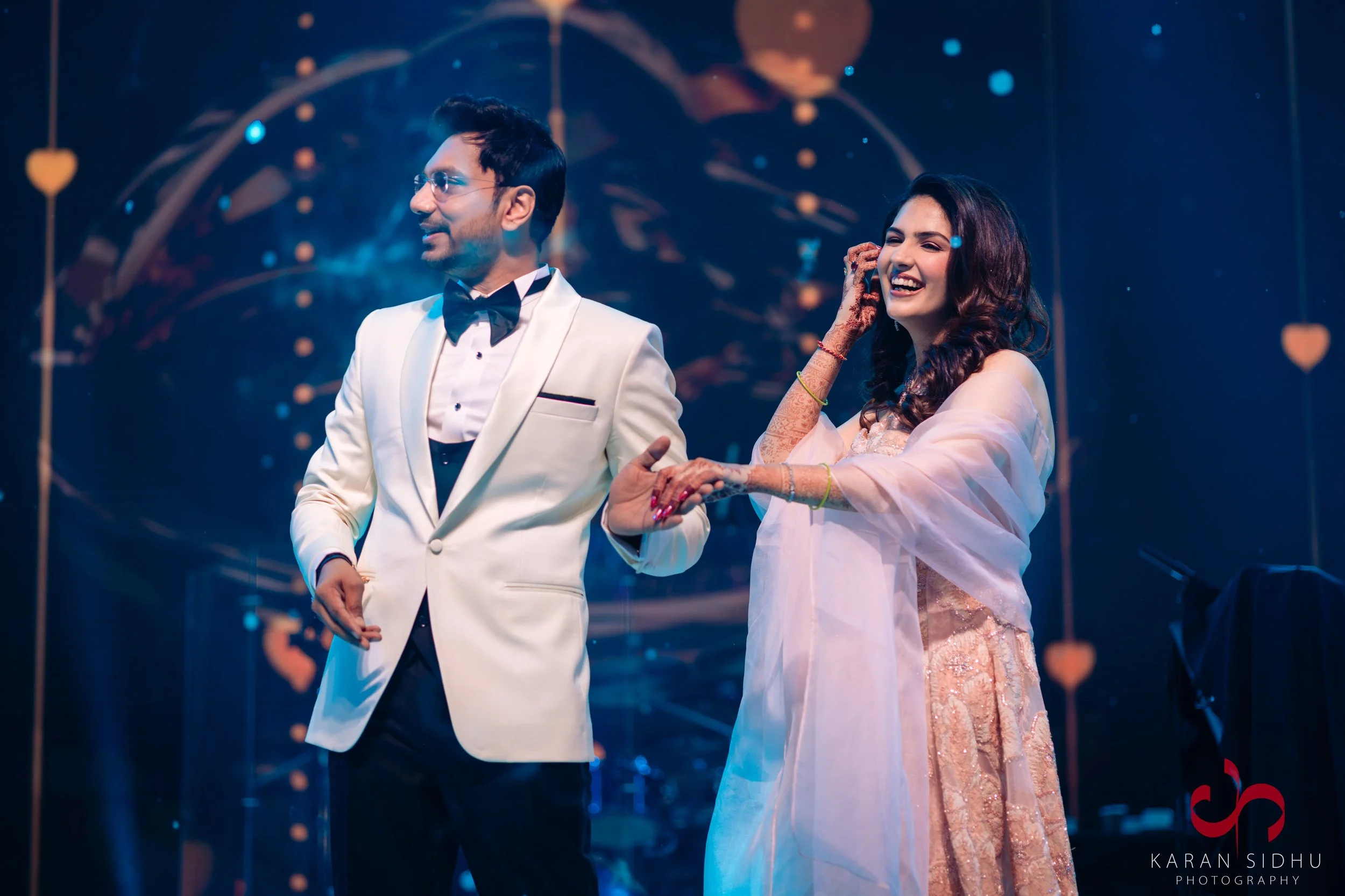 A man in a white tuxedo and a woman in a traditional pink saree dancing together at a celebration event.
