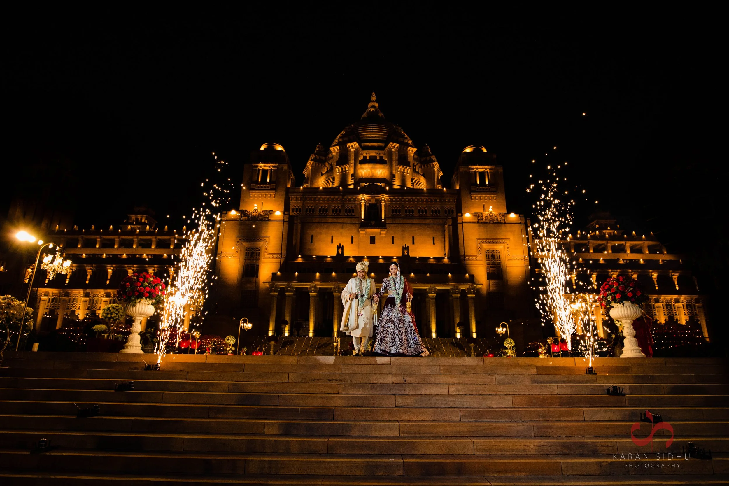 A traditional Indian wedding couple stands on steps in front of a grand, illuminated historic building at night, with fireworks on either side, floral arrangements, and decorative lighting.