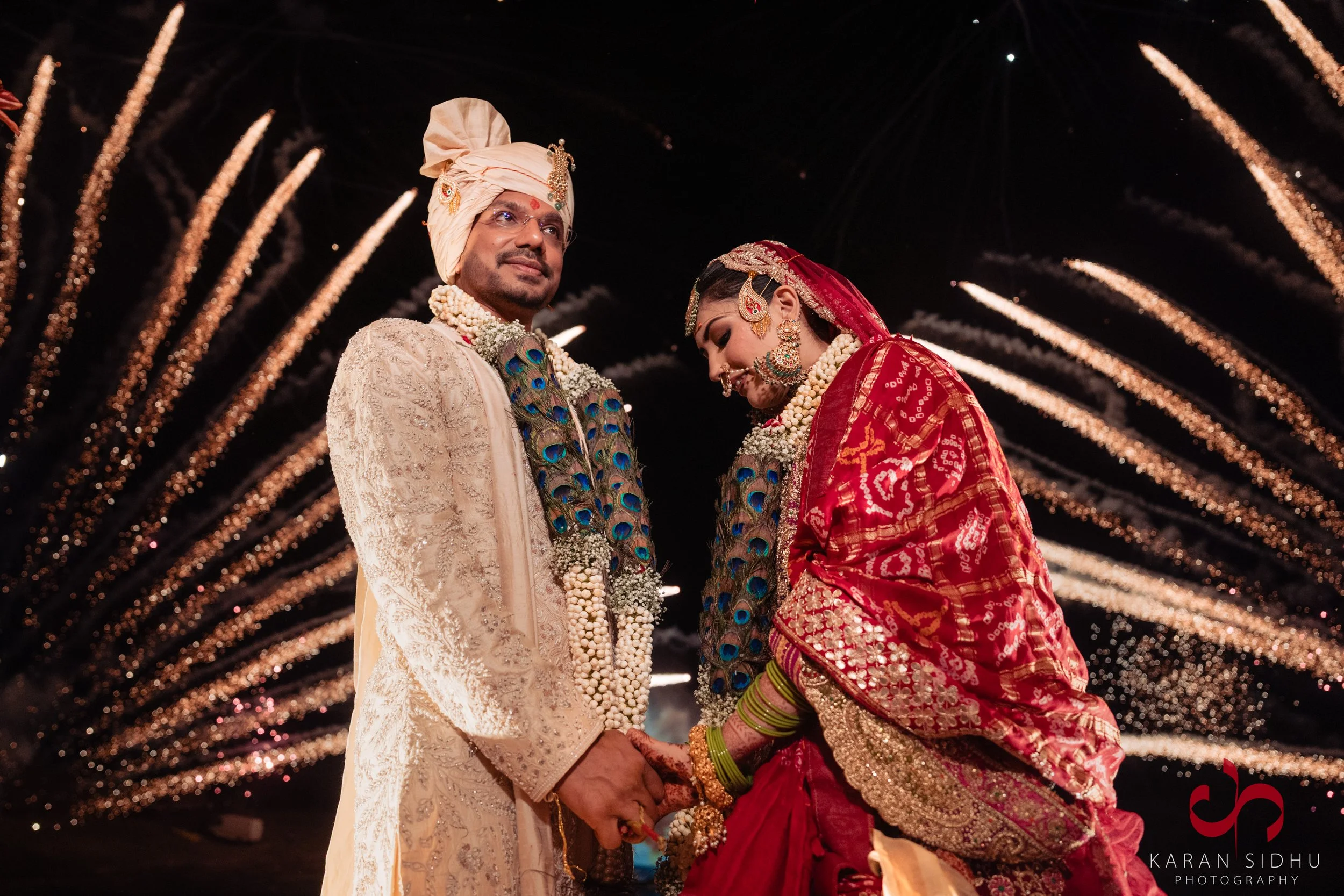 Indian bride and groom holding hands under decorated wedding canopy with fireworks in the background