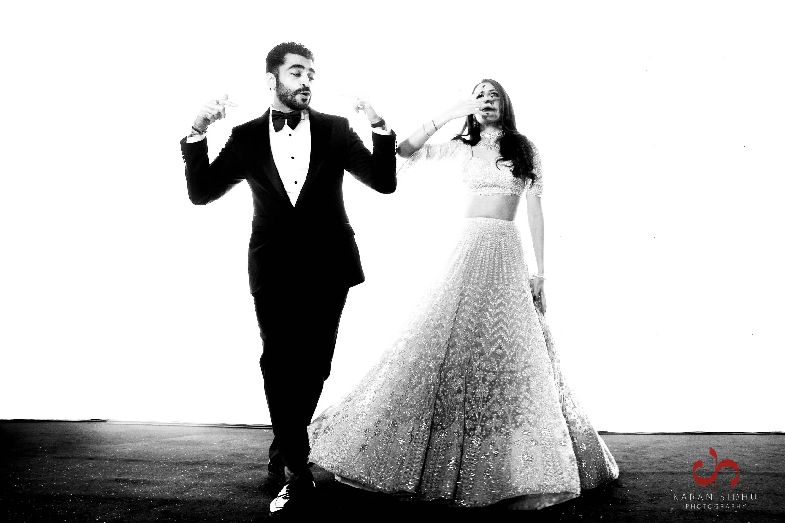 Black and white photo of a man in a tuxedo and a woman in a traditional Indian bridal outfit posing together on a plain backdrop. The man is doing a dance pose with hands near his shoulders, while the woman covers her mouth with her hand. The photographer's logo is in the bottom right corner.