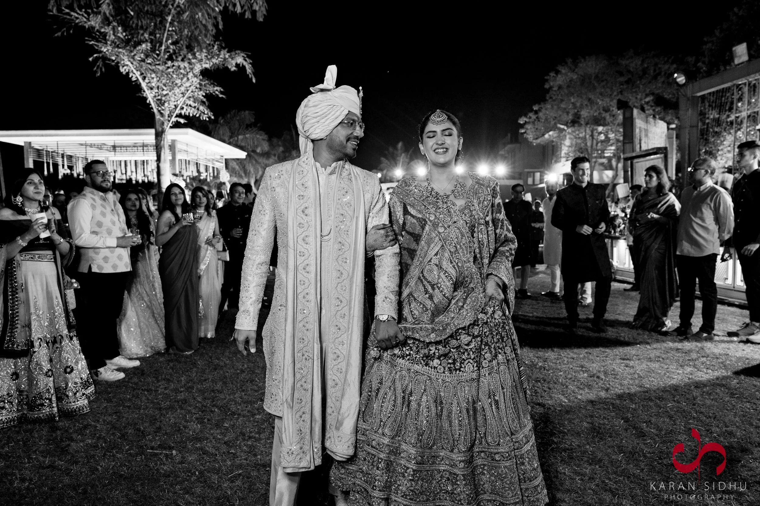 Indian bride and groom walking arm-in-arm at night during wedding celebration, surrounded by guests in traditional attire, outdoors, festive atmosphere.