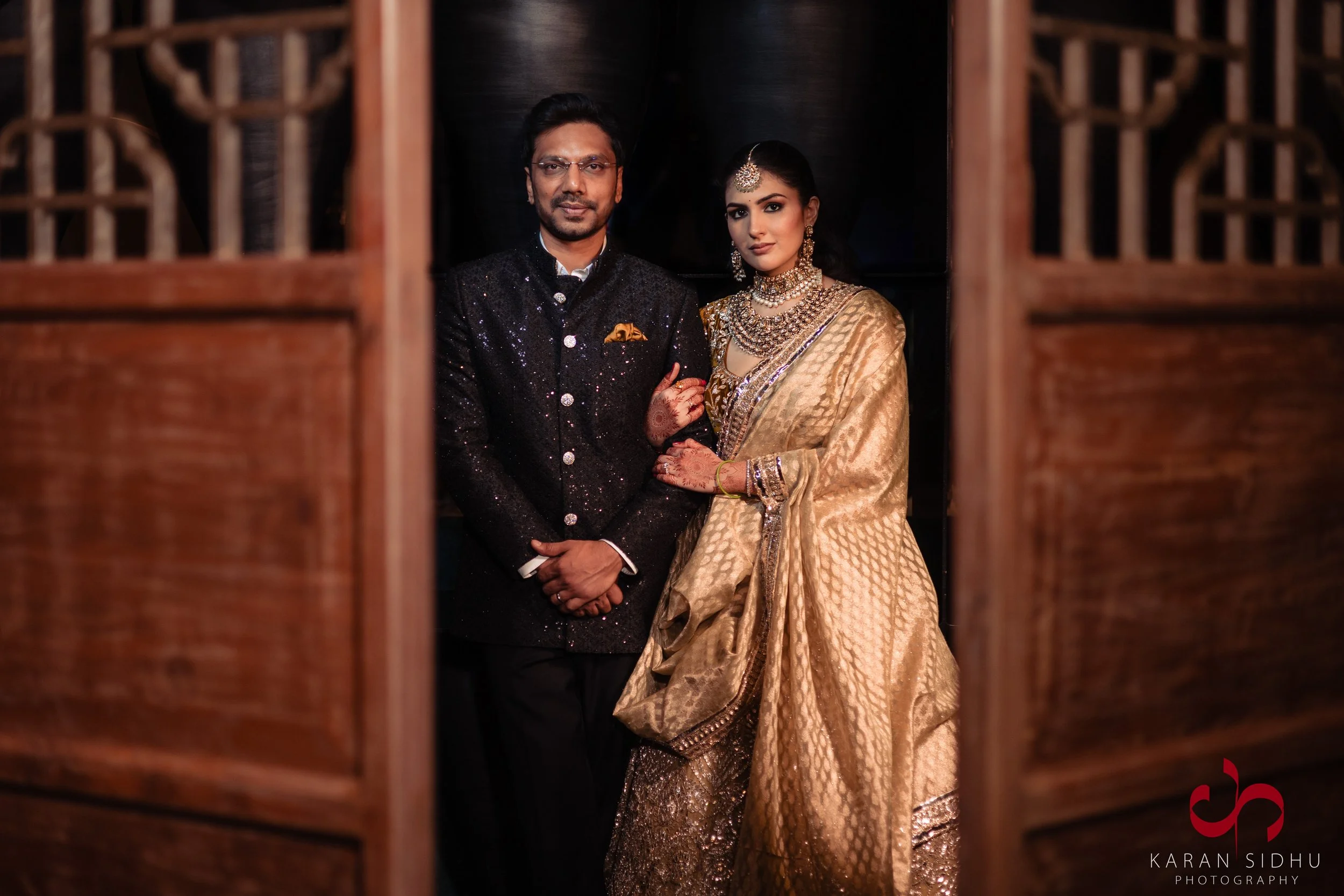 A man and woman dressed in traditional Indian attire standing together in front of a dark background, framed by wooden panels.