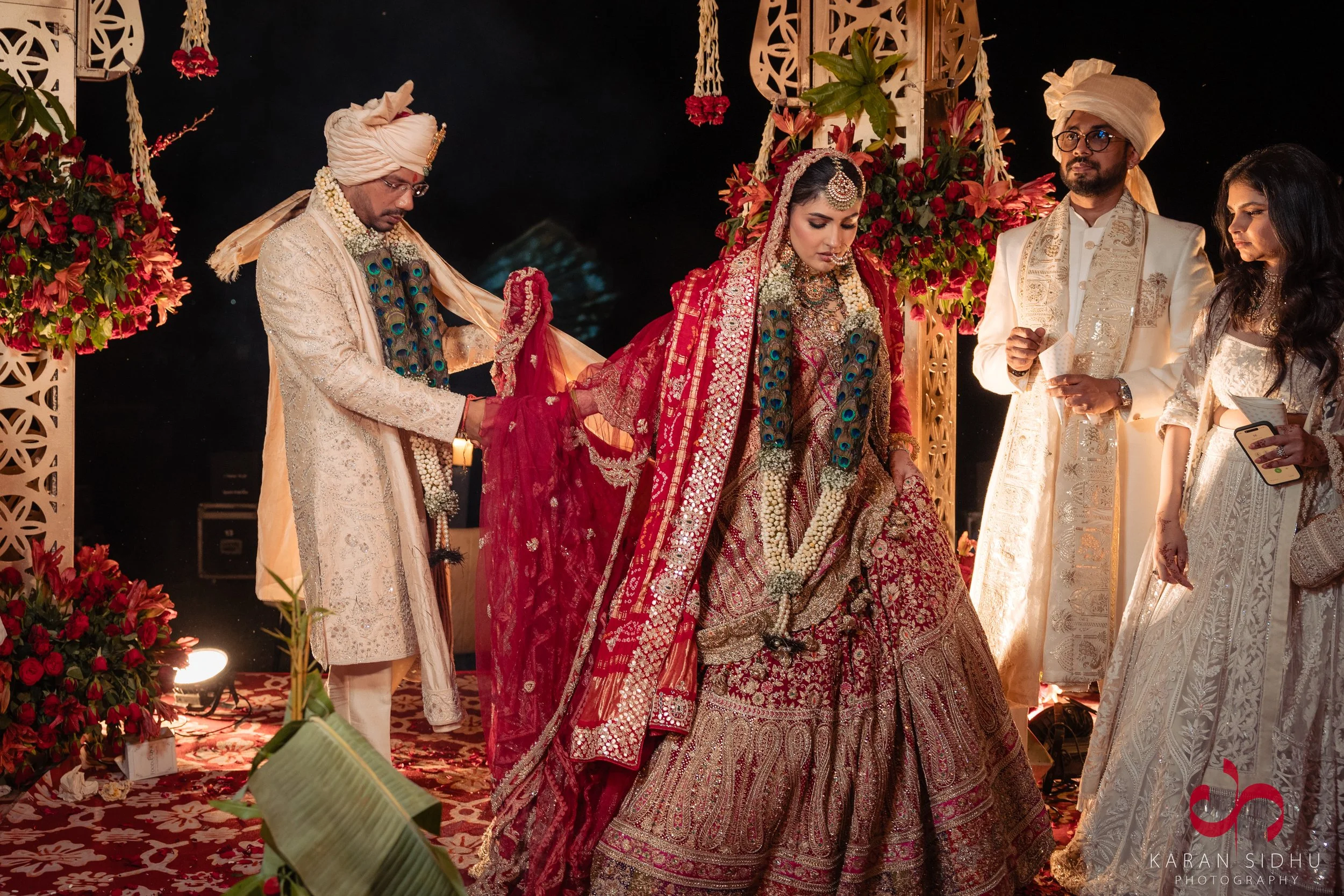 A traditional Indian wedding ceremony with a bride in a red bridal saree and jewelry, standing with assistance, surrounded by ornate floral decorations and family members.