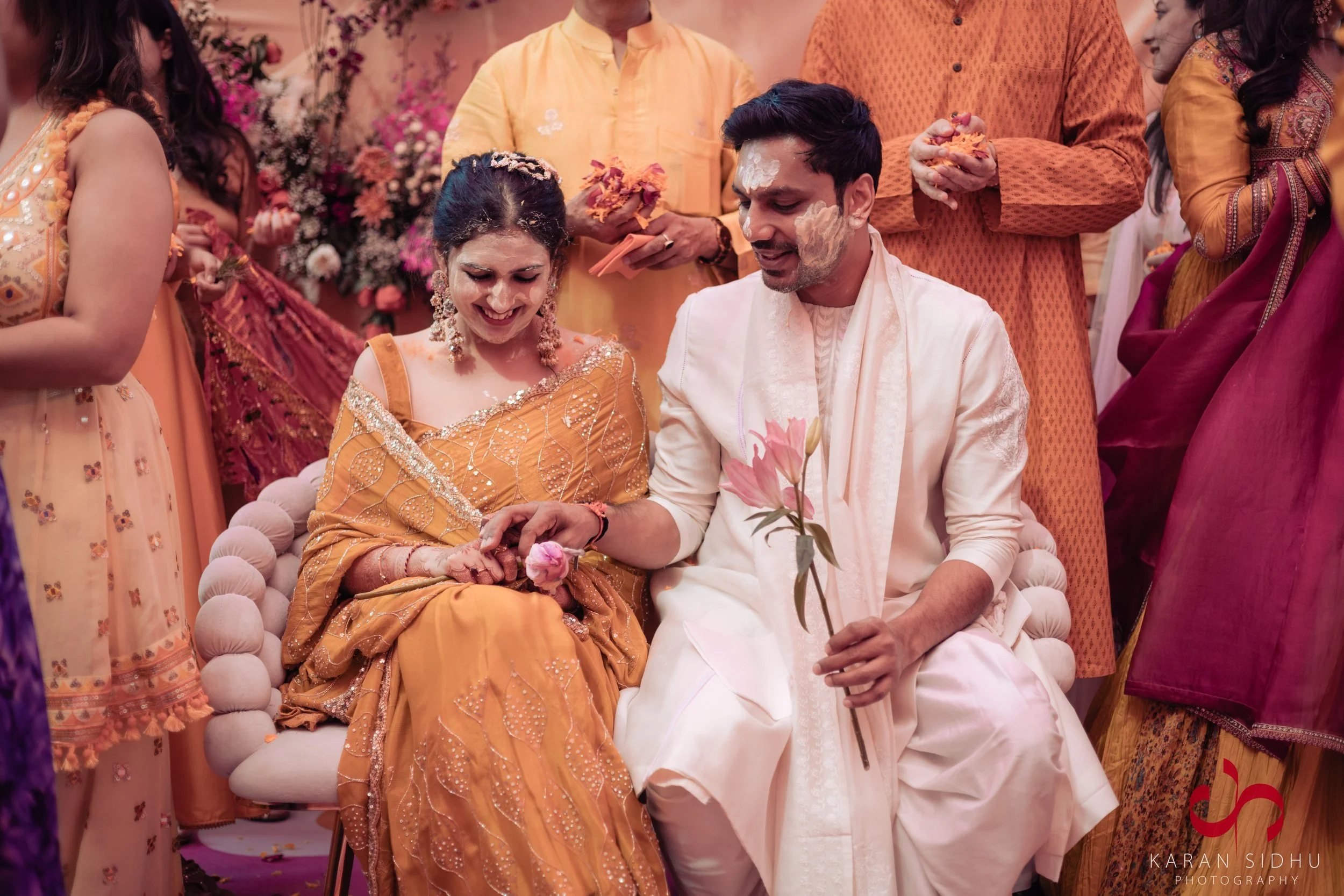 A couple during a traditional Indian wedding ceremony, surrounded by family and friends, with flower petals and various colors of attire, all celebrating in a festive setting.