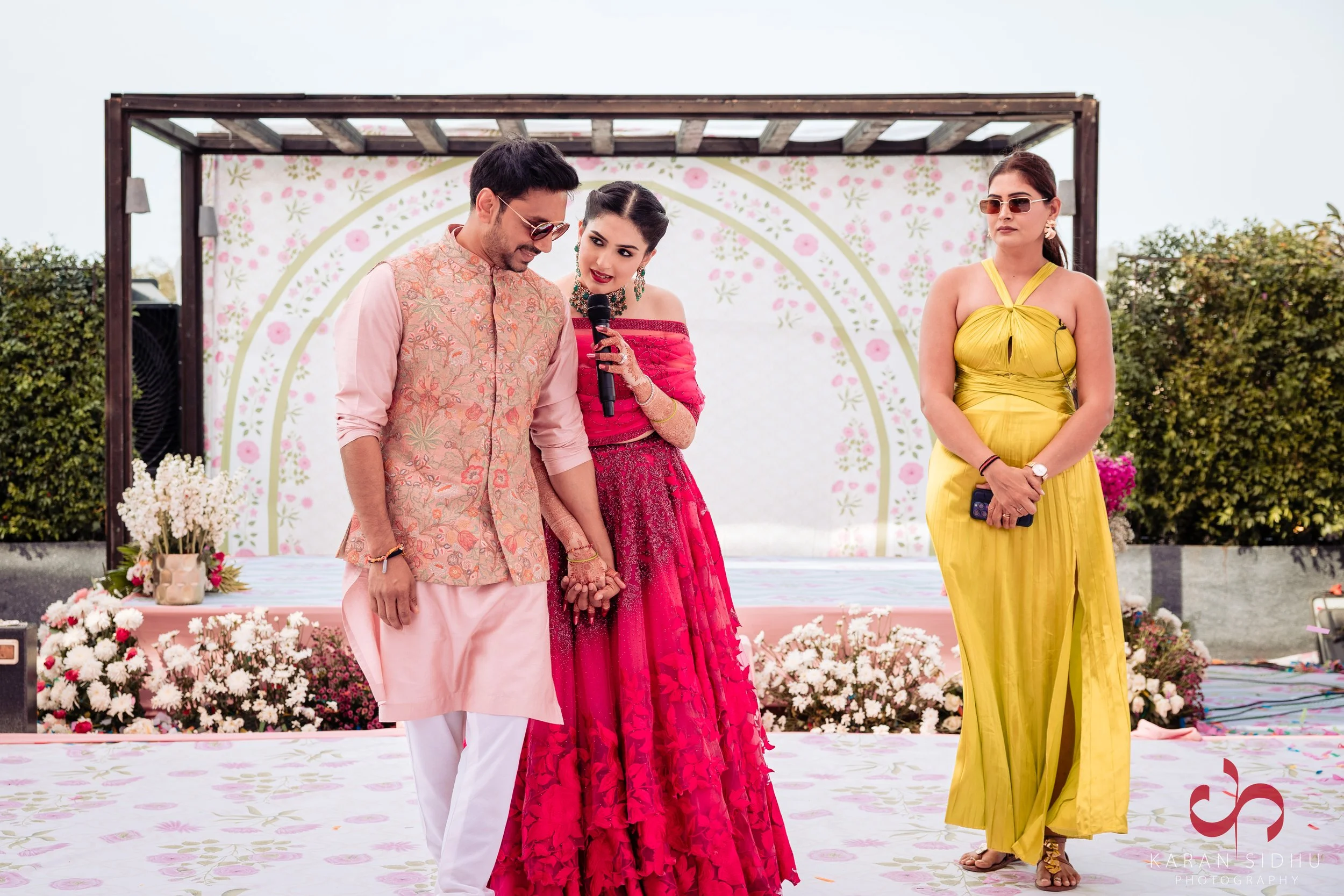 A couple dressed in traditional Indian wedding attire holding hands on a decorated outdoor stage, with a woman in a yellow dress standing nearby.