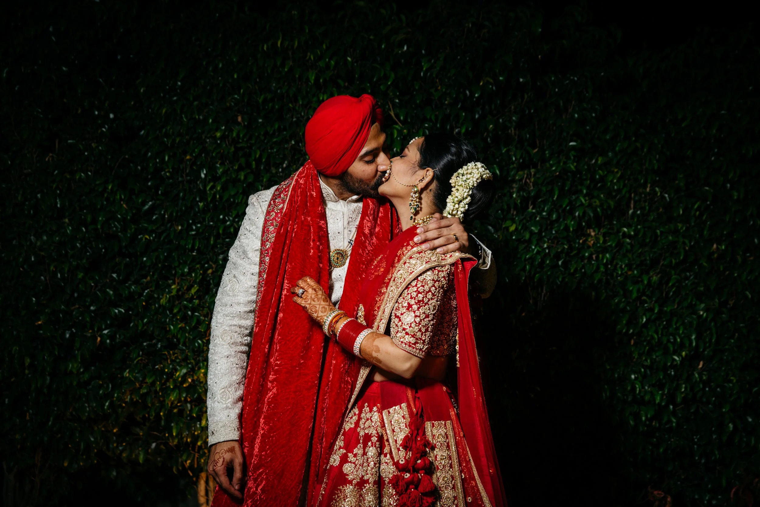 An Indian couple dressed in traditional wedding attire sharing a kiss outdoors at night, with green foliage in the background.