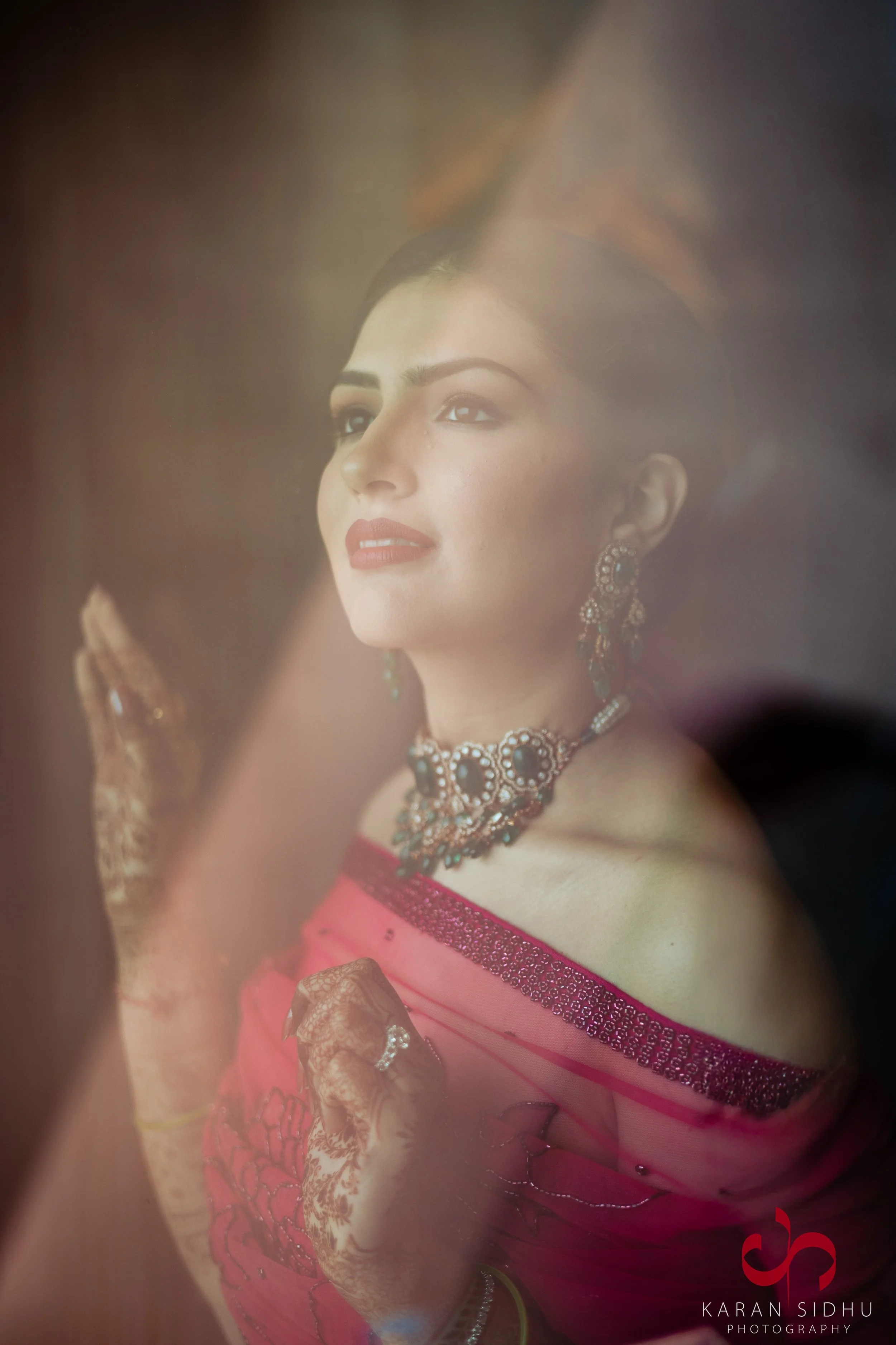 A woman with traditional Indian jewelry and Mehndi art on her hands, dressed in a pink saree, looking through a window with a dreamy expression.
