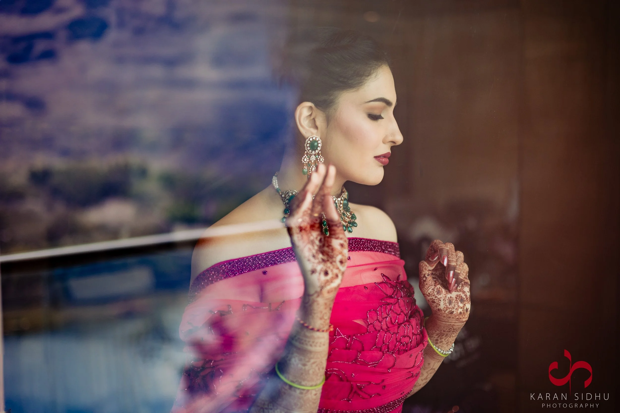 A woman dressed in traditional Indian attire with henna on her hands, jewelry, and makeup, looking out a window with her hand gently touching the glass.