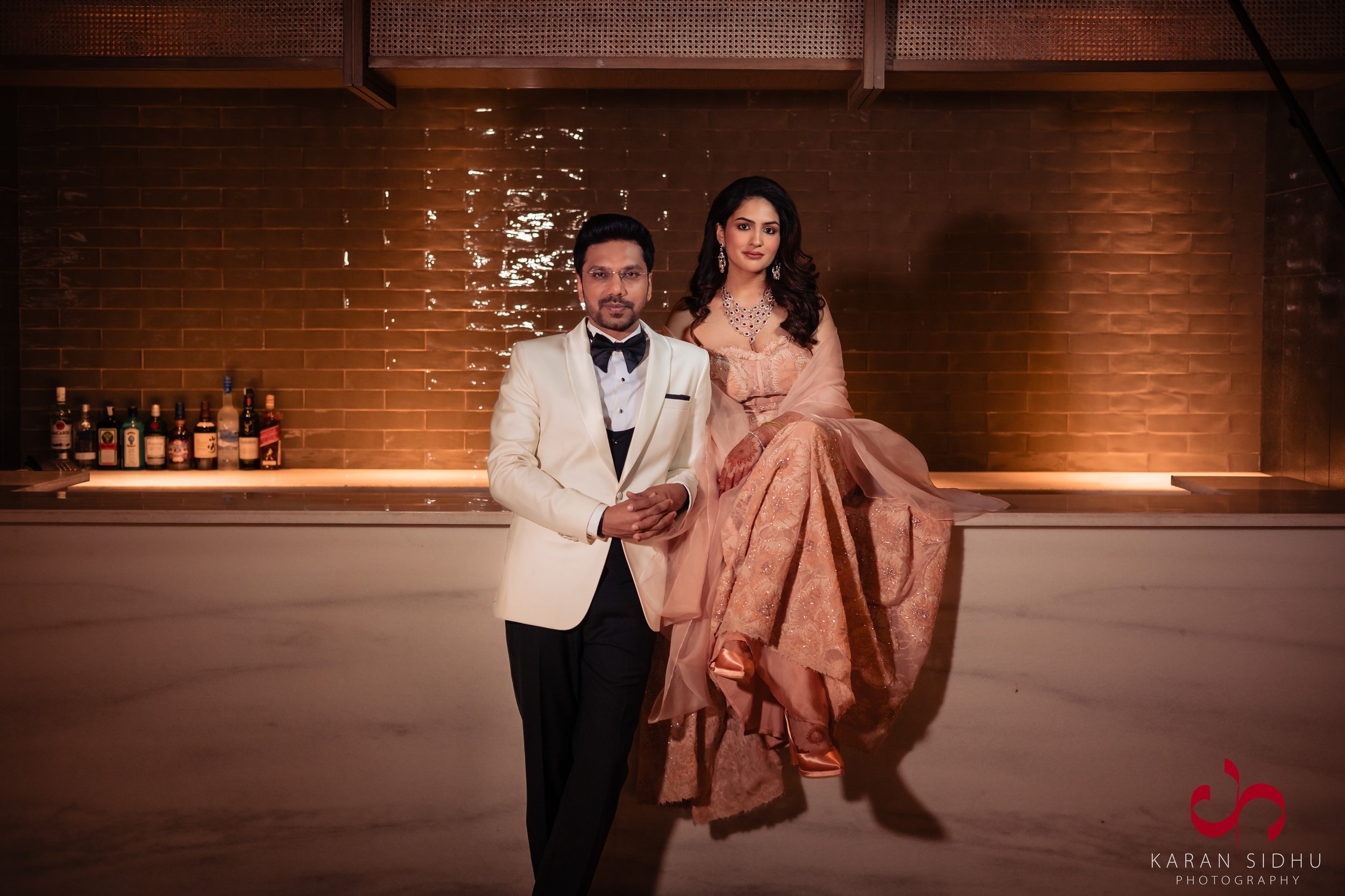 A man in a white tuxedo with a black bow tie standing next to a woman dressed in an elegant peach-colored gown, seated on a bar counter with bottles of alcohol in the background, in a dimly-lit, stylish setting.