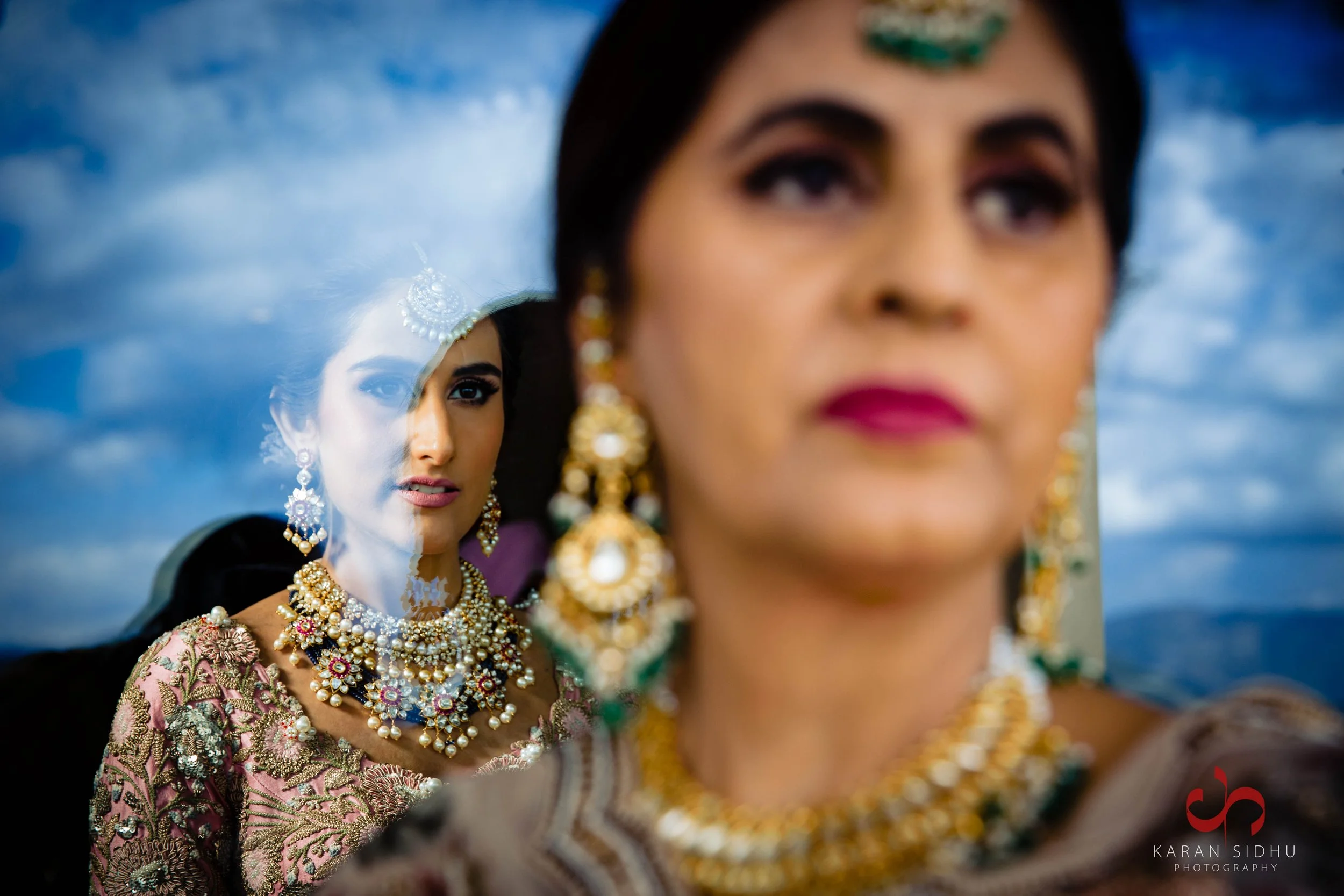 A woman wearing traditional Indian jewelry and attire, viewed through a mirror showing her reflection. The background features clouds in the sky.
