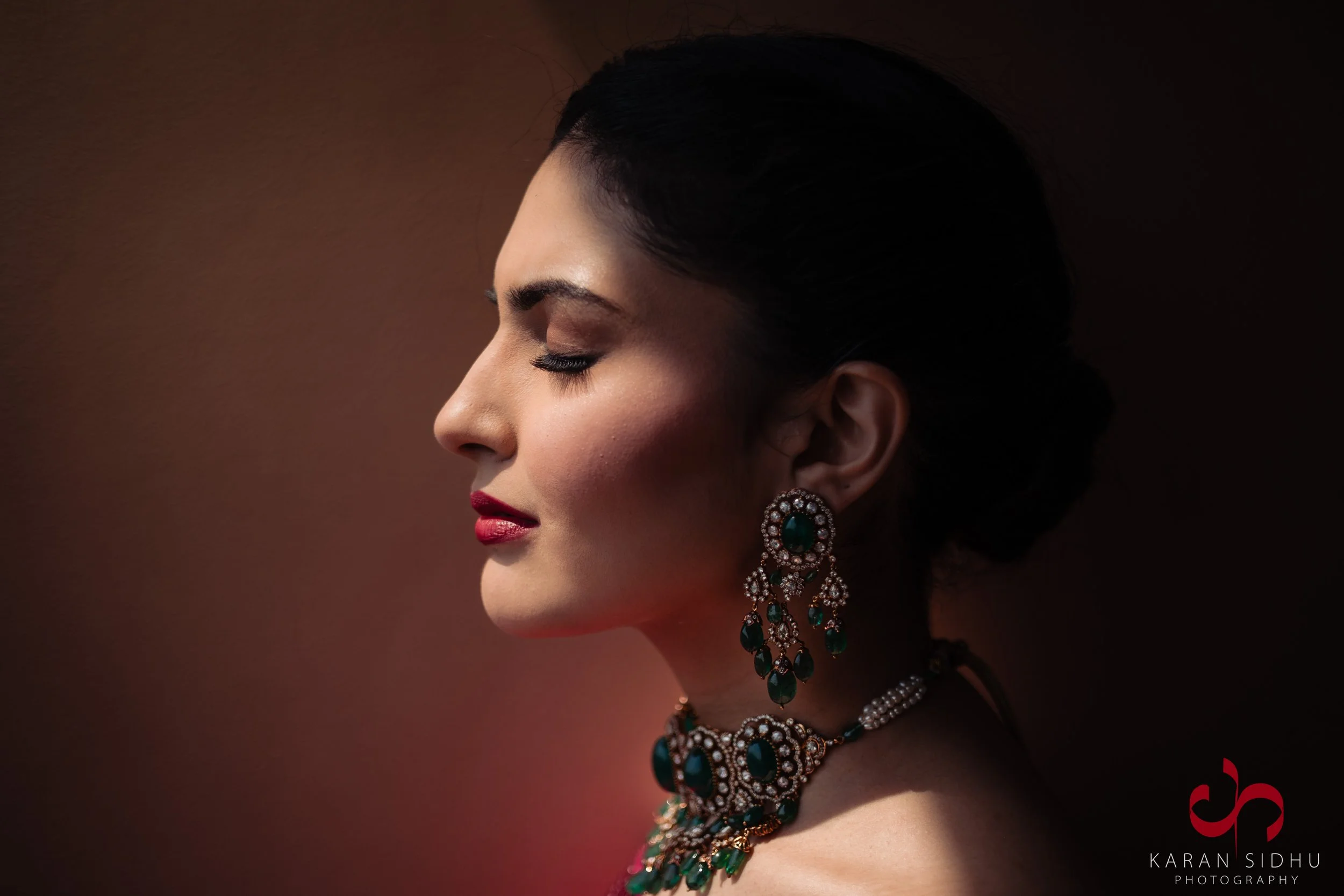 Profile of a woman with closed eyes, wearing ornate earrings and a matching necklace, with a dark blurred background.