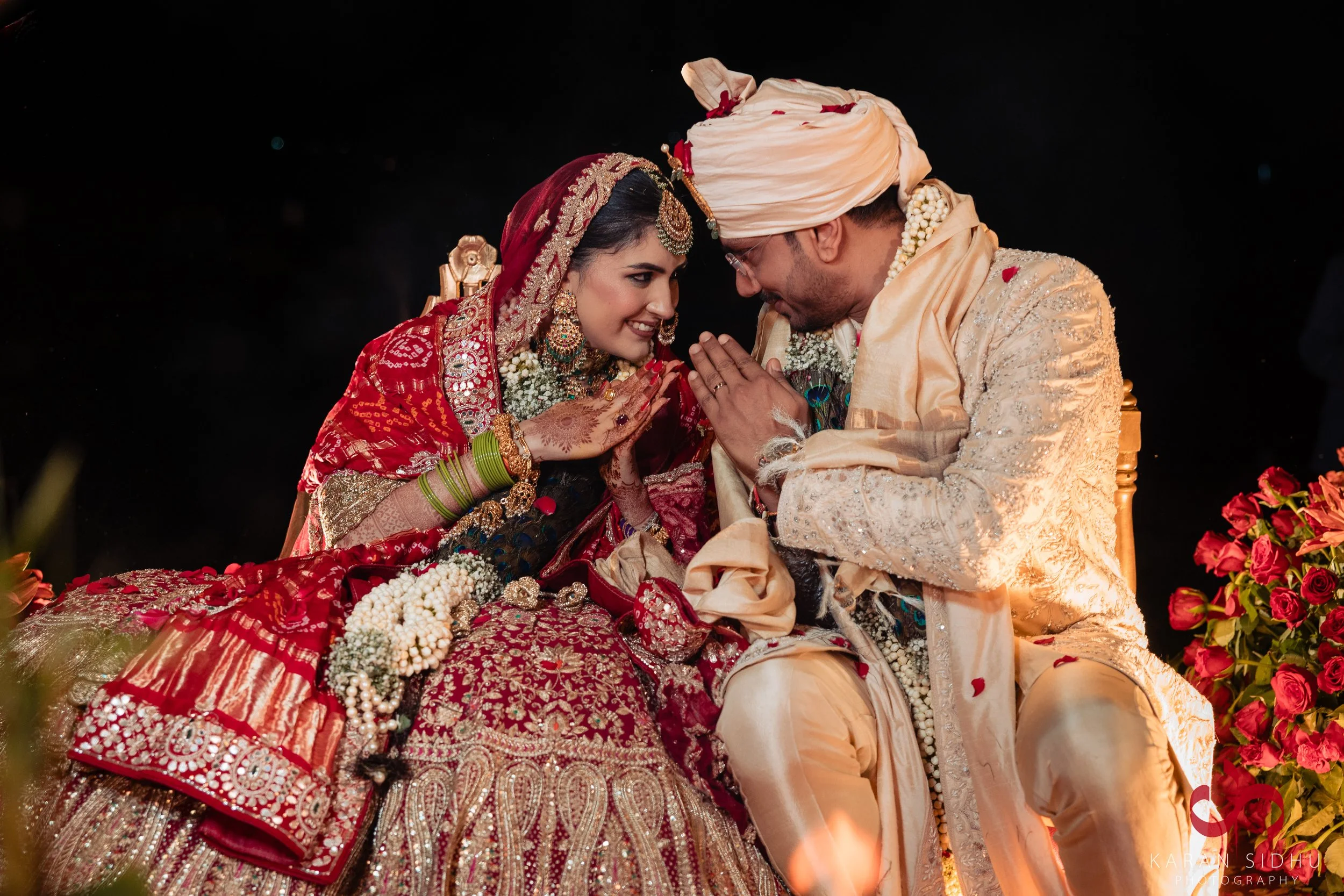 A bride and groom in traditional Indian wedding attire sharing a moment, sitting close and touching foreheads, with flowers and decorative elements around them.