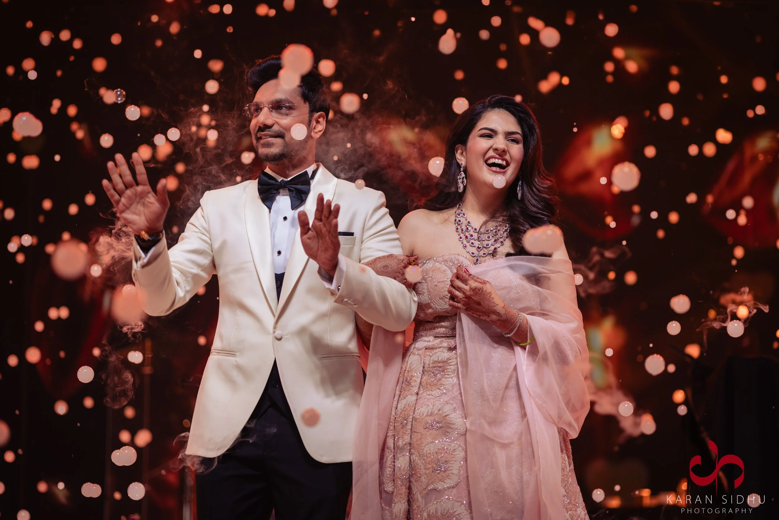 A couple dressed in formal wedding attire celebrating at a wedding reception with sparklers and bokeh lights in the background. The man is wearing a white tuxedo with a black bow tie, and the woman is wearing a pink bridal gown with jewelry. They are
