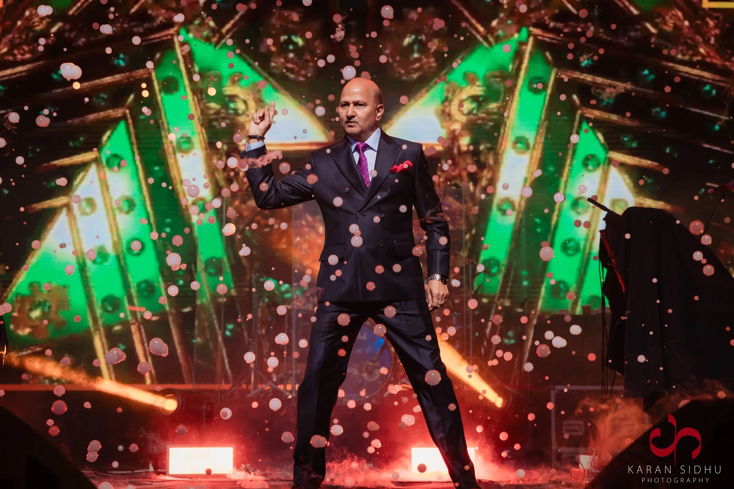 A man in a dark double-breasted suit with a pink tie and matching pocket square is performing on stage with a microphone in the background. He is standing with one arm raised, surrounded by colorful lighting, confetti, and smoke effects. The stage ha