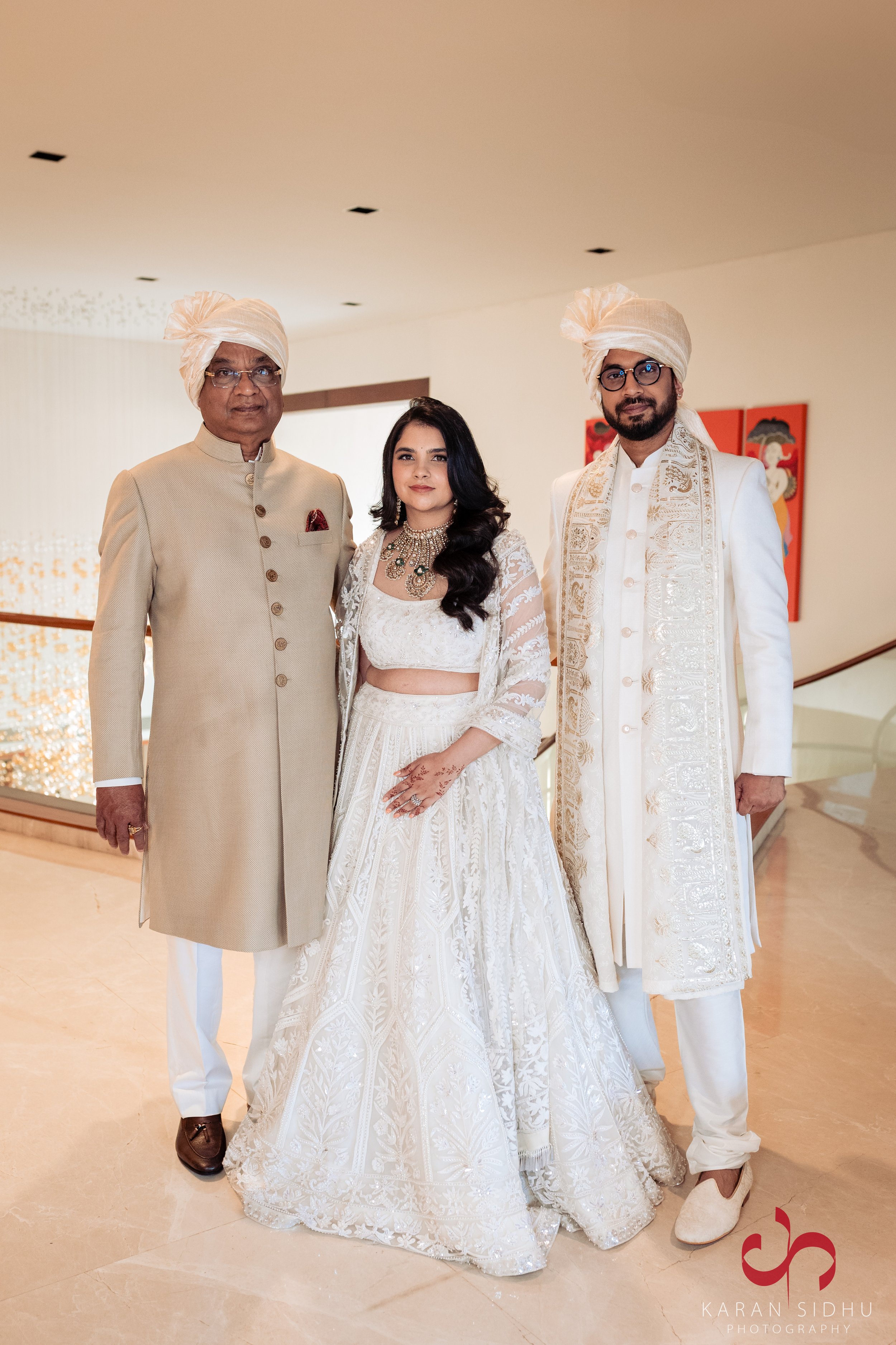 A group of three people dressed in traditional Indian wedding attire, standing indoors. The man on the left is wearing a beige sherwani with brown shoes, a cream turban, and glasses. The woman in the middle is wearing a white embroidered bridal lehen