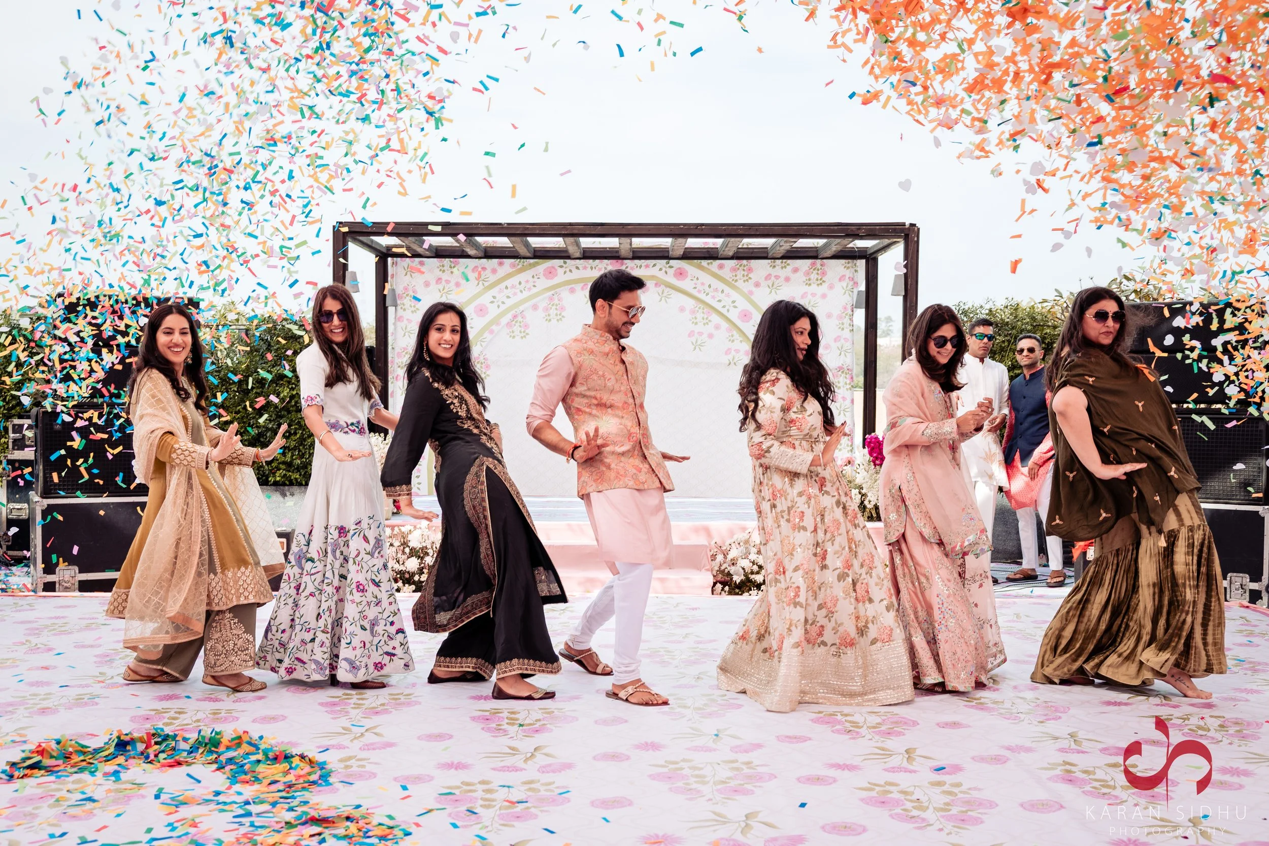 People celebrating at an outdoor event with colorful confetti falling, dressed in traditional Indian attire, standing on decorated stage with a floral backdrop.