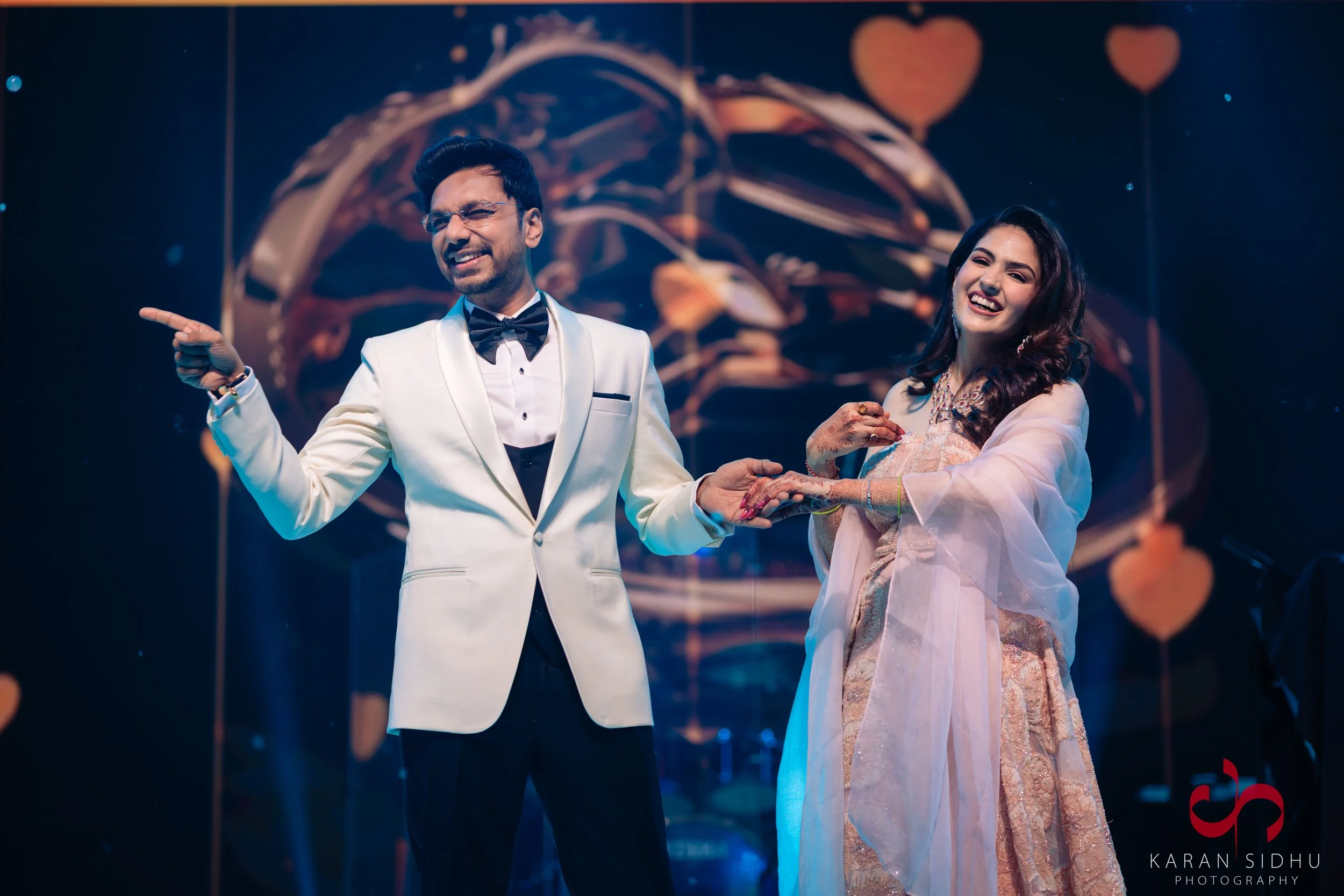 A man in a white tuxedo with a black bow tie and glasses, and a woman in a gold and pink traditional dress, smiling and holding hands on a stage decorated with heart-shaped lanterns in the background.