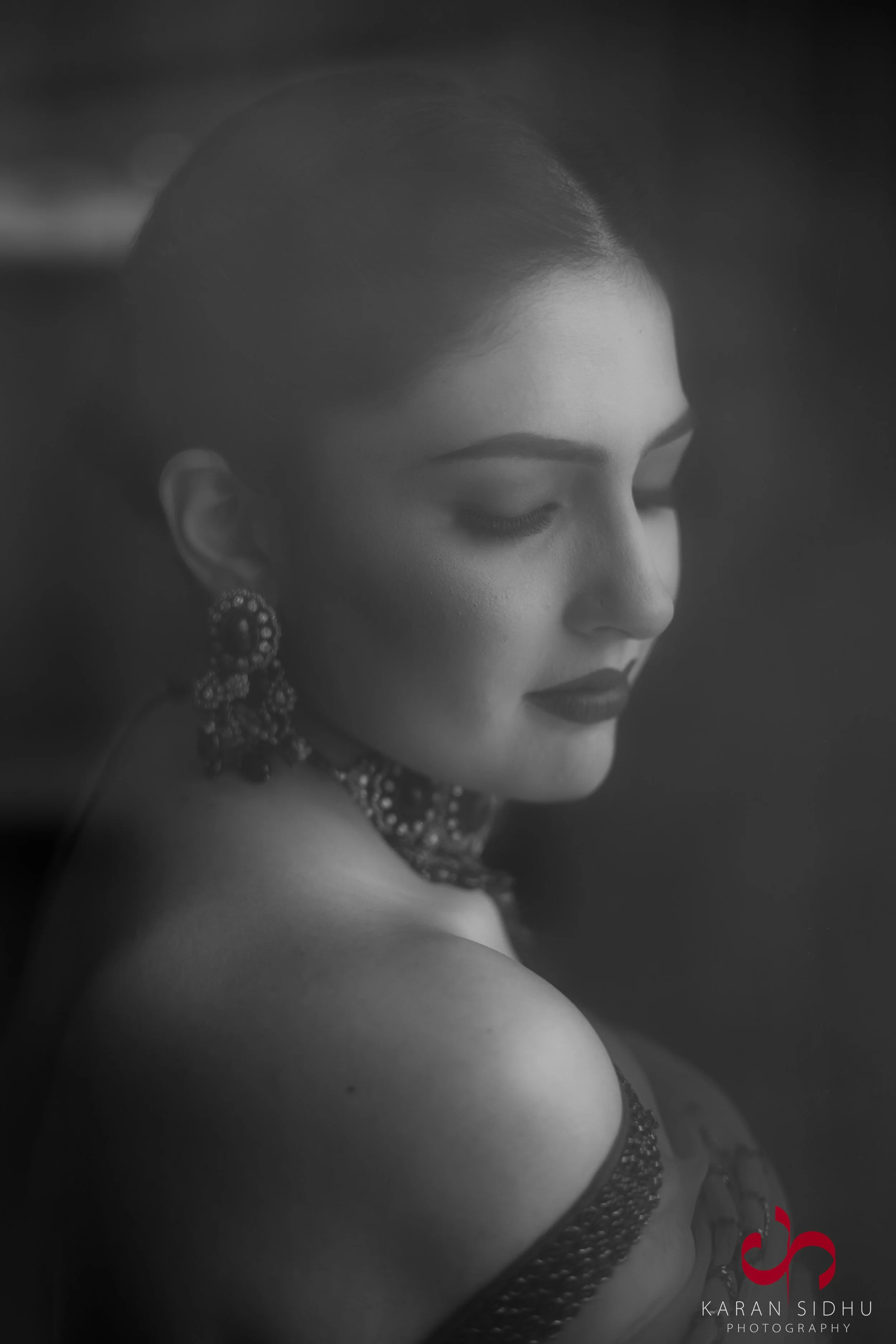Black and white portrait of a woman with closed eyes and dark lipstick, wearing intricate earrings and a necklace, with a soft focus and artistic portrait style.