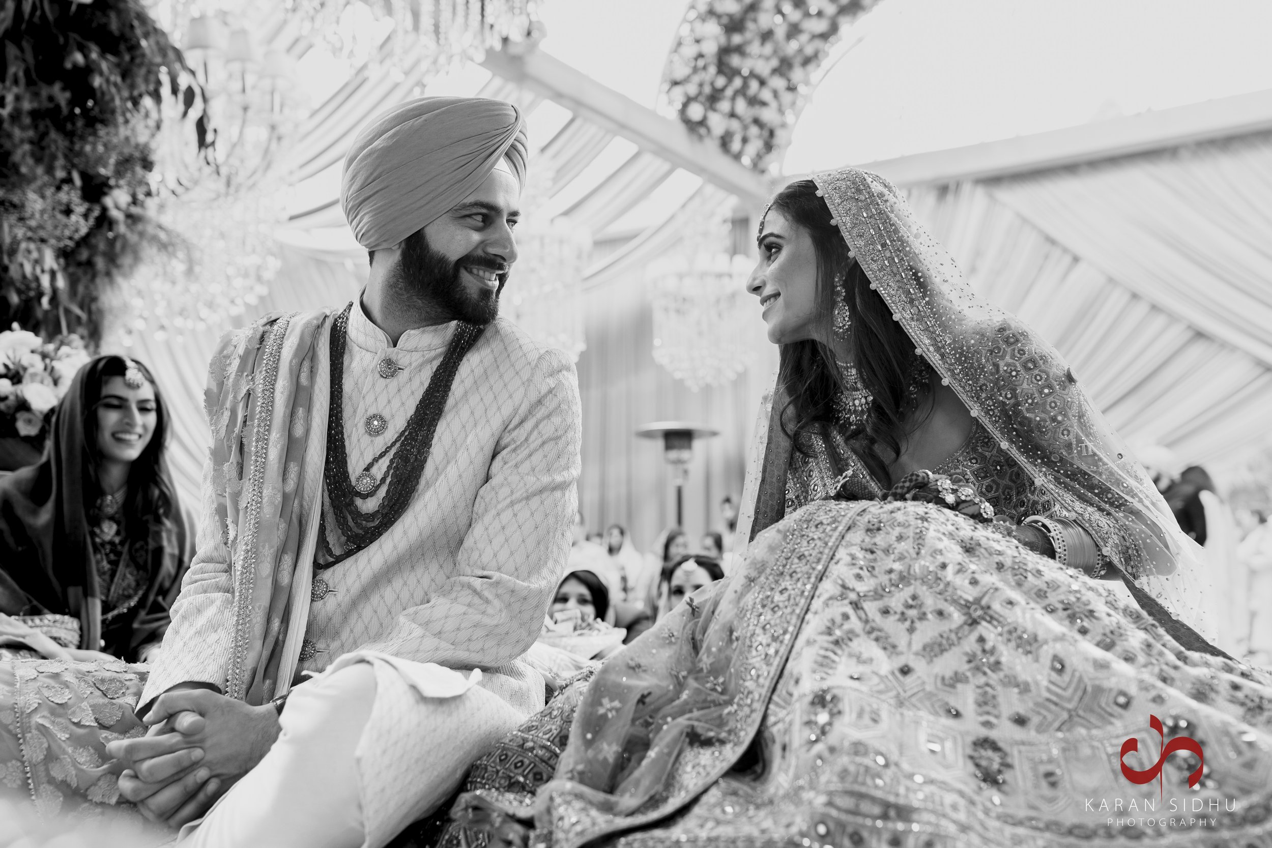 A bride and groom sit together during an Indian wedding ceremony, dressed in traditional attire, smiling at each other, with wedding guests in the background.
