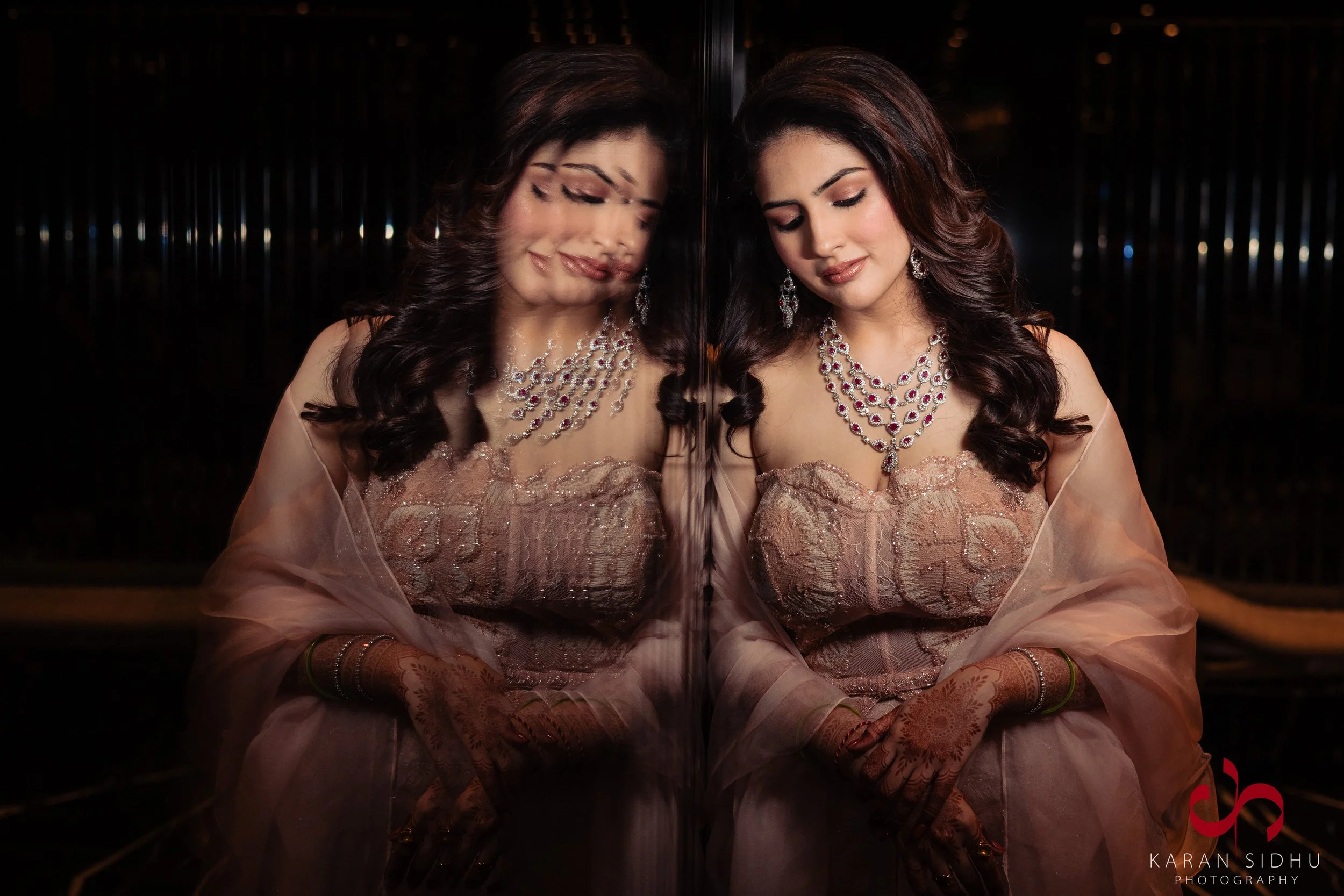 A woman in a beige saree and heavy jewelry leaning against a mirror, reflection shows her face with closed eyes and a gentle smile.