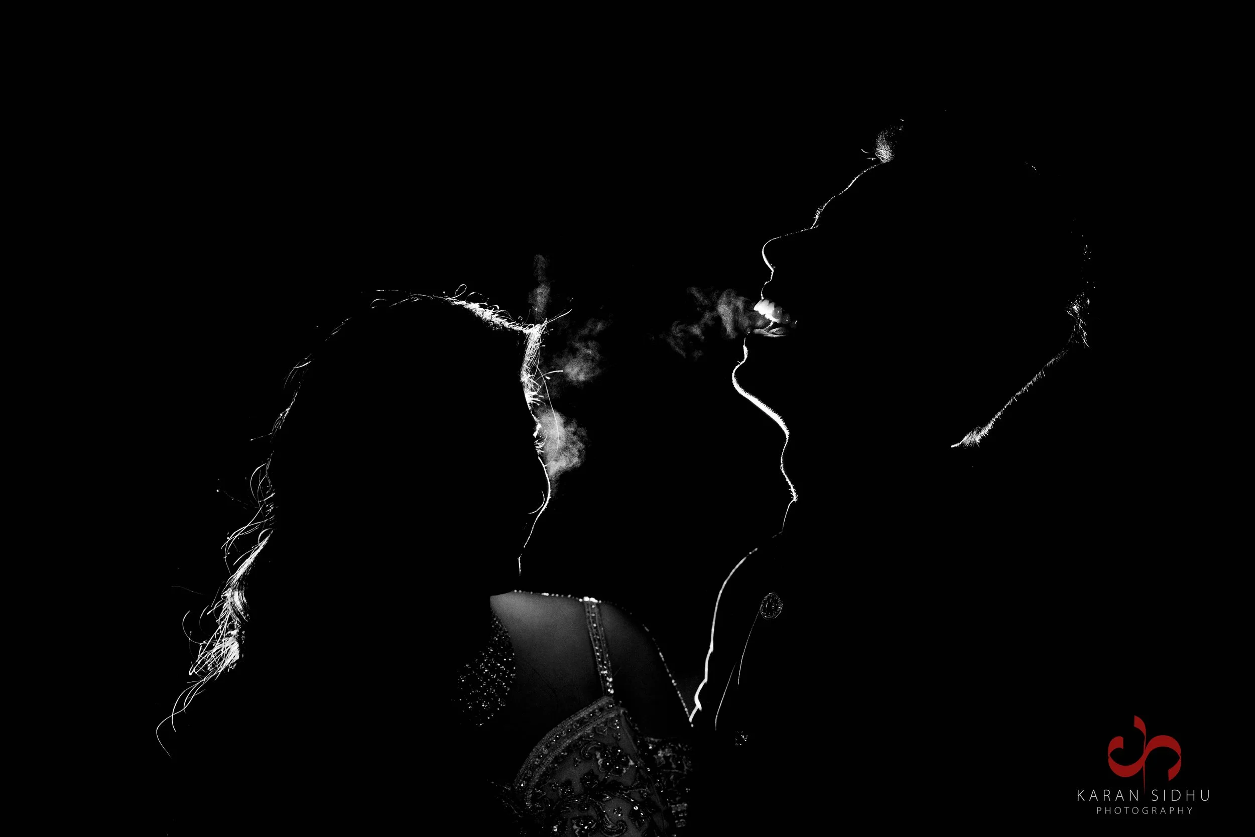 Silhouette of a couple facing each other with smoke or vapor, highlighting their profiles, against a black background.