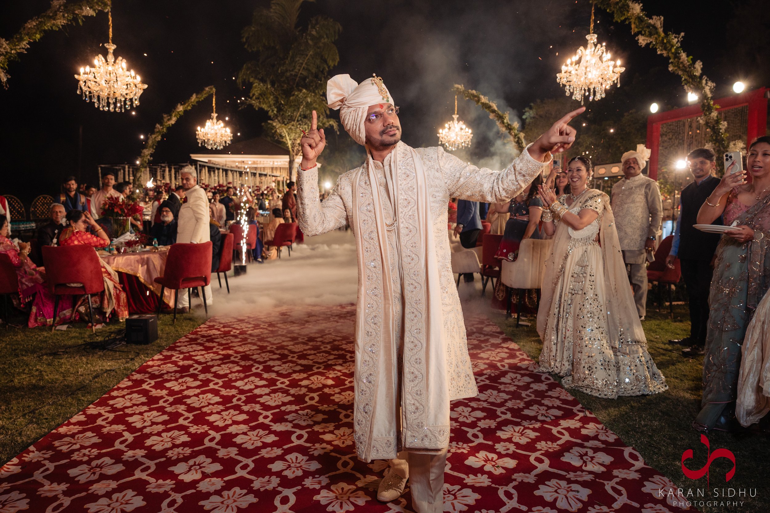 A groom dressed in traditional Indian wedding attire, including a cream-colored sherwani and turban, stands on a decorated red carpet at an outdoor night wedding reception. He is making a dance move with both arms raised and pointing upward. Guests i