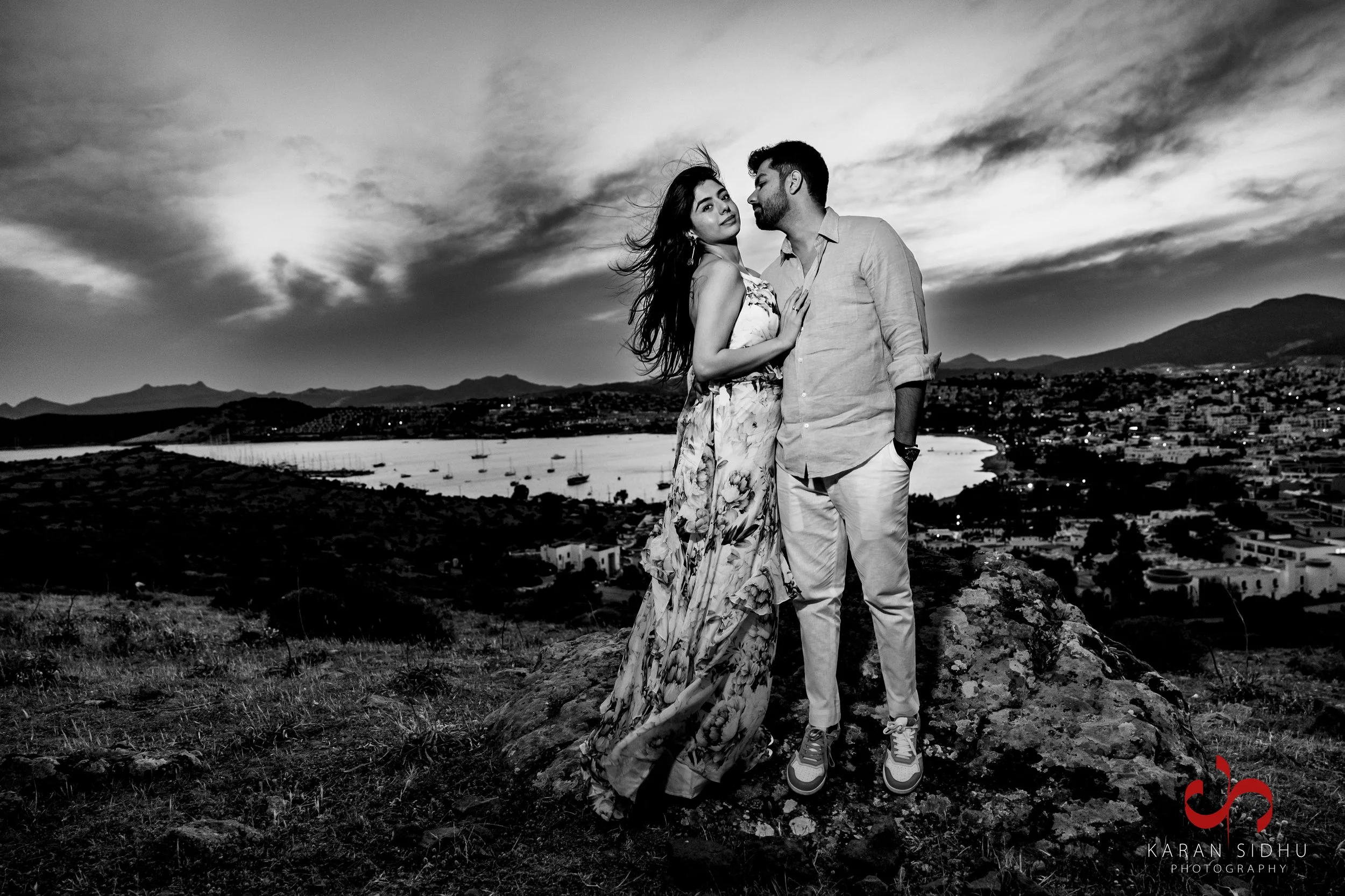A black and white photo of a couple standing on a rocky hilltop at sunset with a city view and mountains in the background. The woman wears a floral dress, and the man wears a casual shirt and pants.