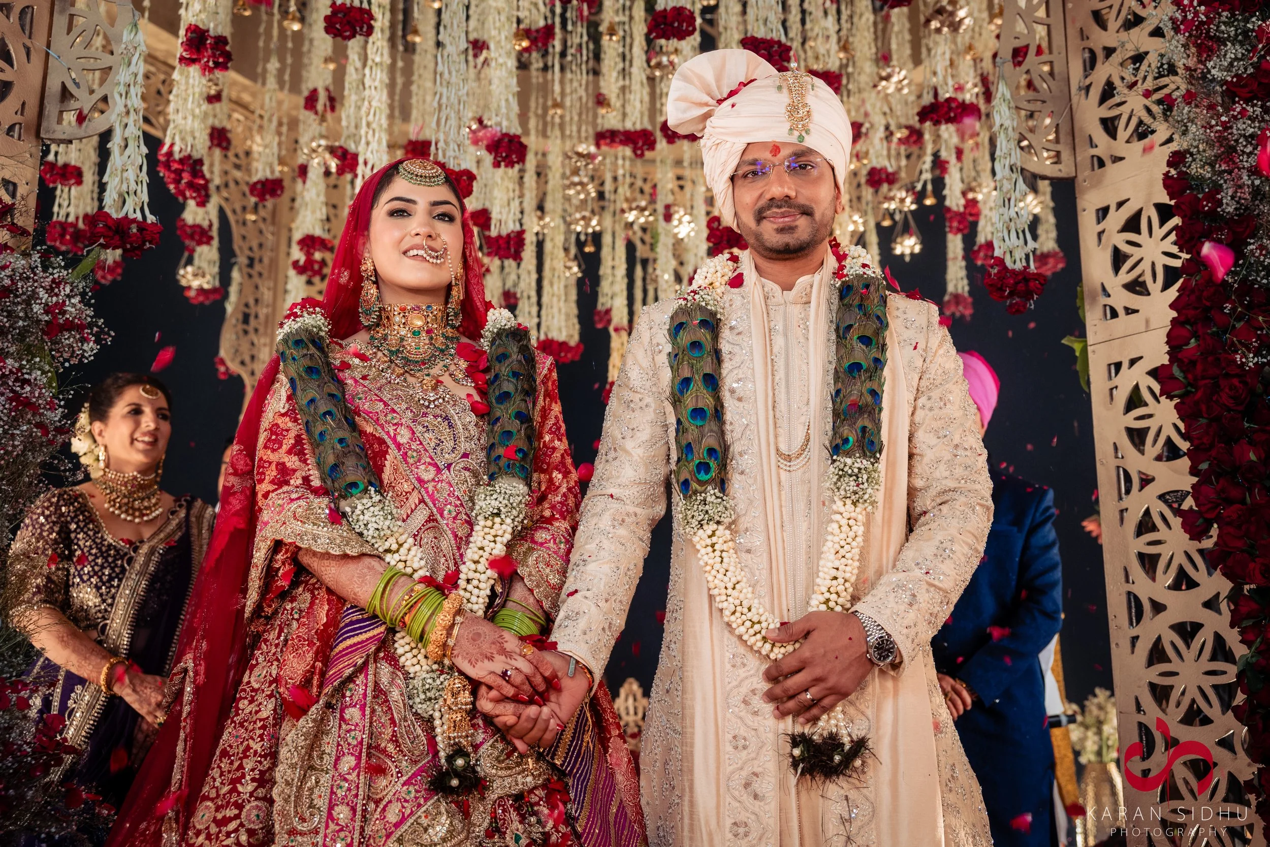 A newlywed Indian couple at their wedding ceremony, holding hands and surrounded by floral decorations. The bride is dressed in a red and gold lehenga with traditional jewelry, including a nose ring and green bangles. The groom is in a cream-colored 