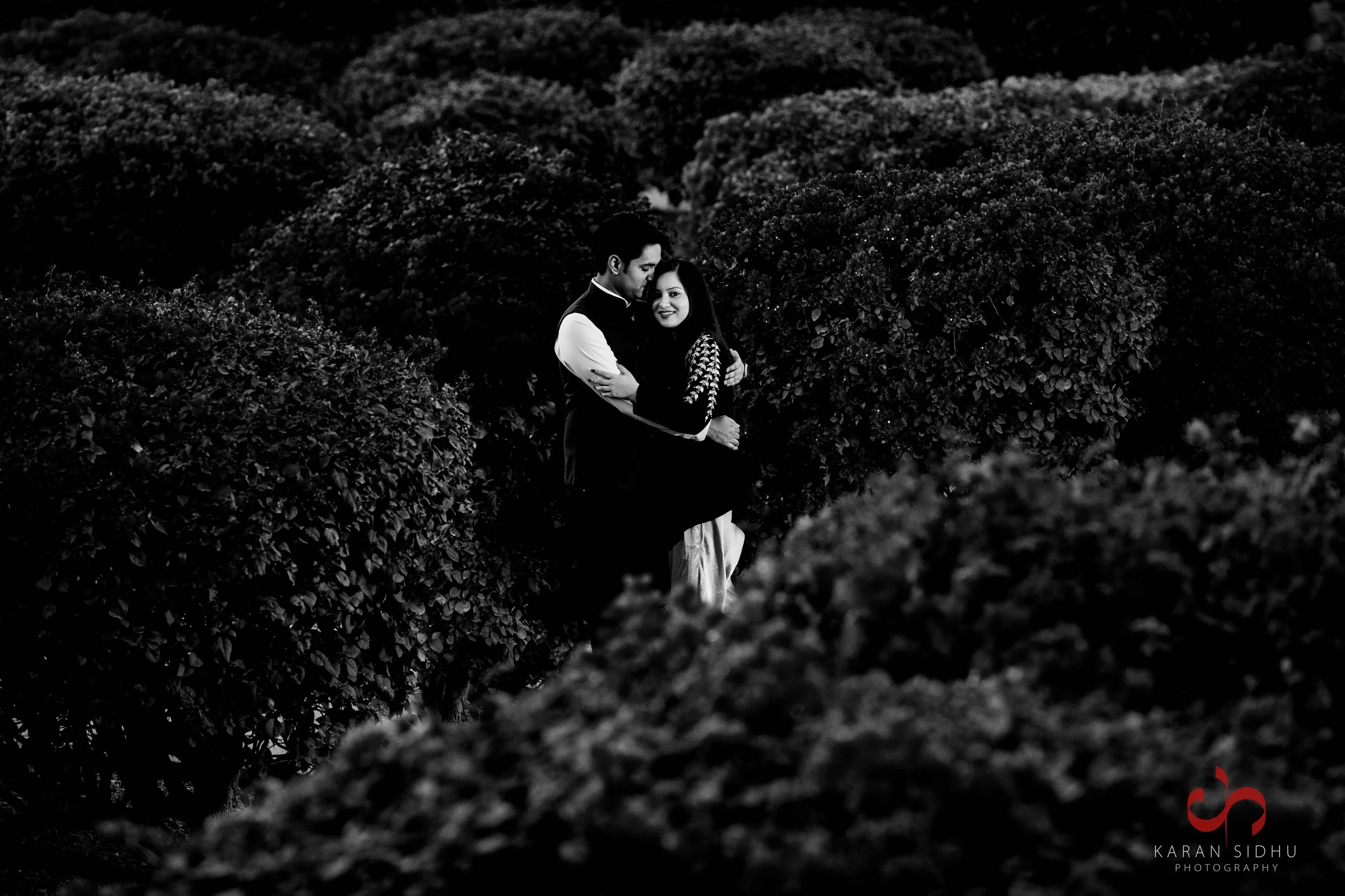 A black-and-white photo of a couple embracing among dense, rounded bushes. The man is wearing a suit with a white shirt, and the woman is in a dark dress with intricate sleeve details. They are smiling and standing close together.