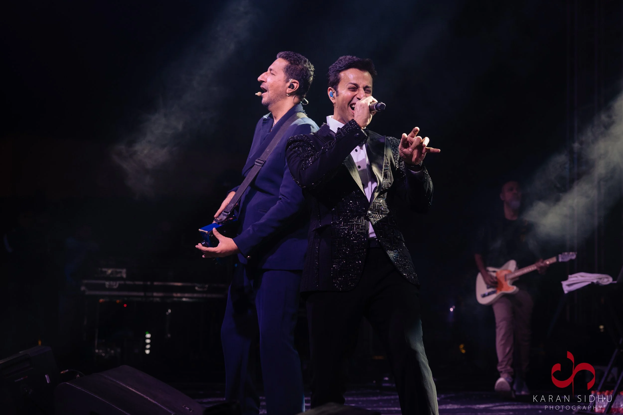 Two male performers singing and playing guitar on stage with a dark background, bright stage lights, and a third musician in the background with a guitar.