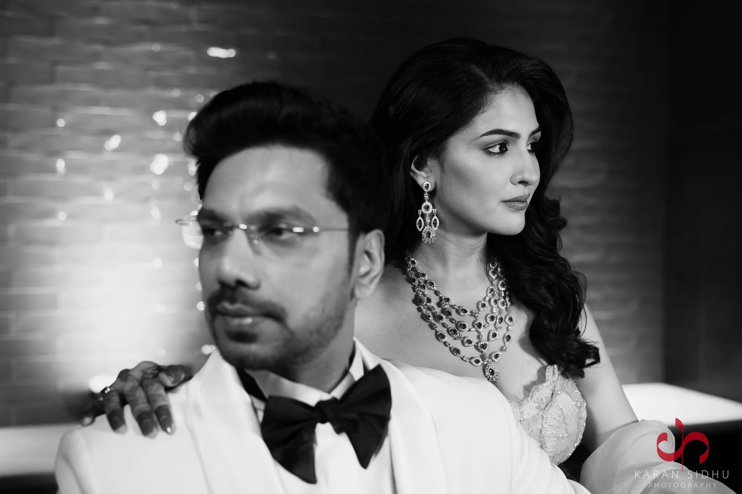 A black and white photo of a man and woman in formal attire, with the man in a white tuxedo and bow tie, and the woman dressed in an elegant gown with jewelry, both looking away from each other.