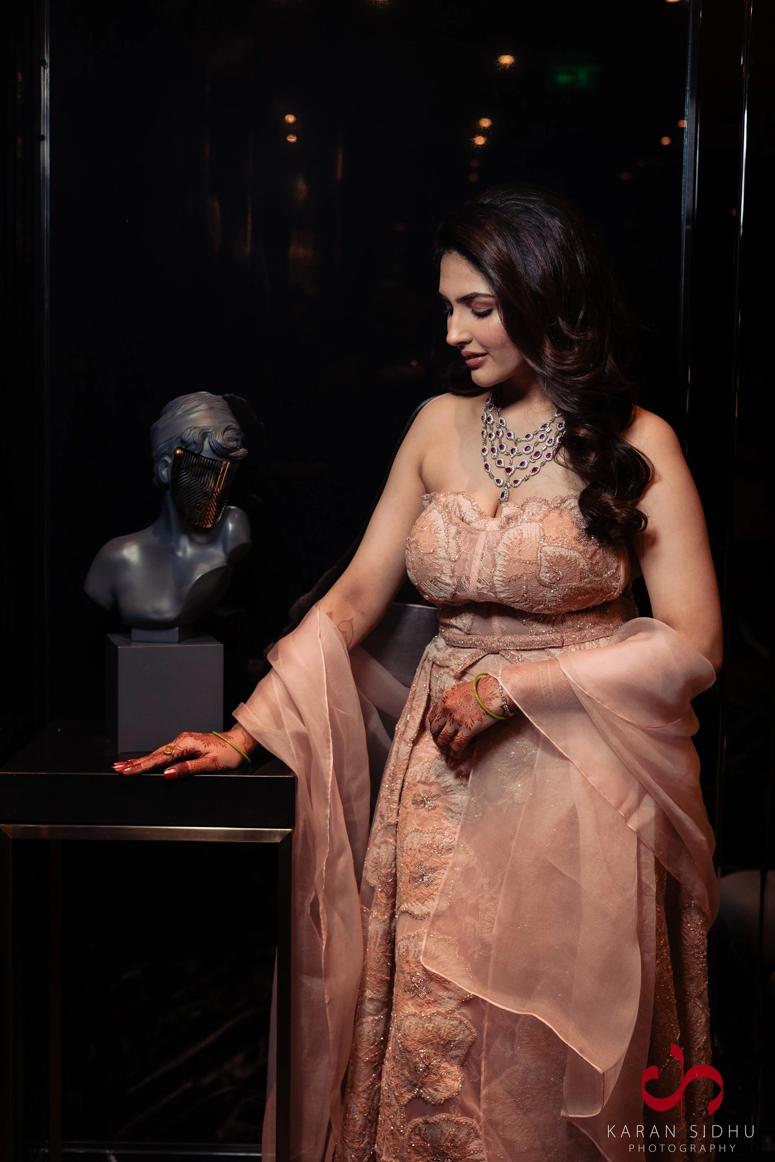 A woman in an elegant peach-colored saree and jewelry is standing beside a black pedestal with a sculpture of a person's bust with long hair and a faceless face. She is looking down, with her left hand resting on the pedestal and her right hand on he